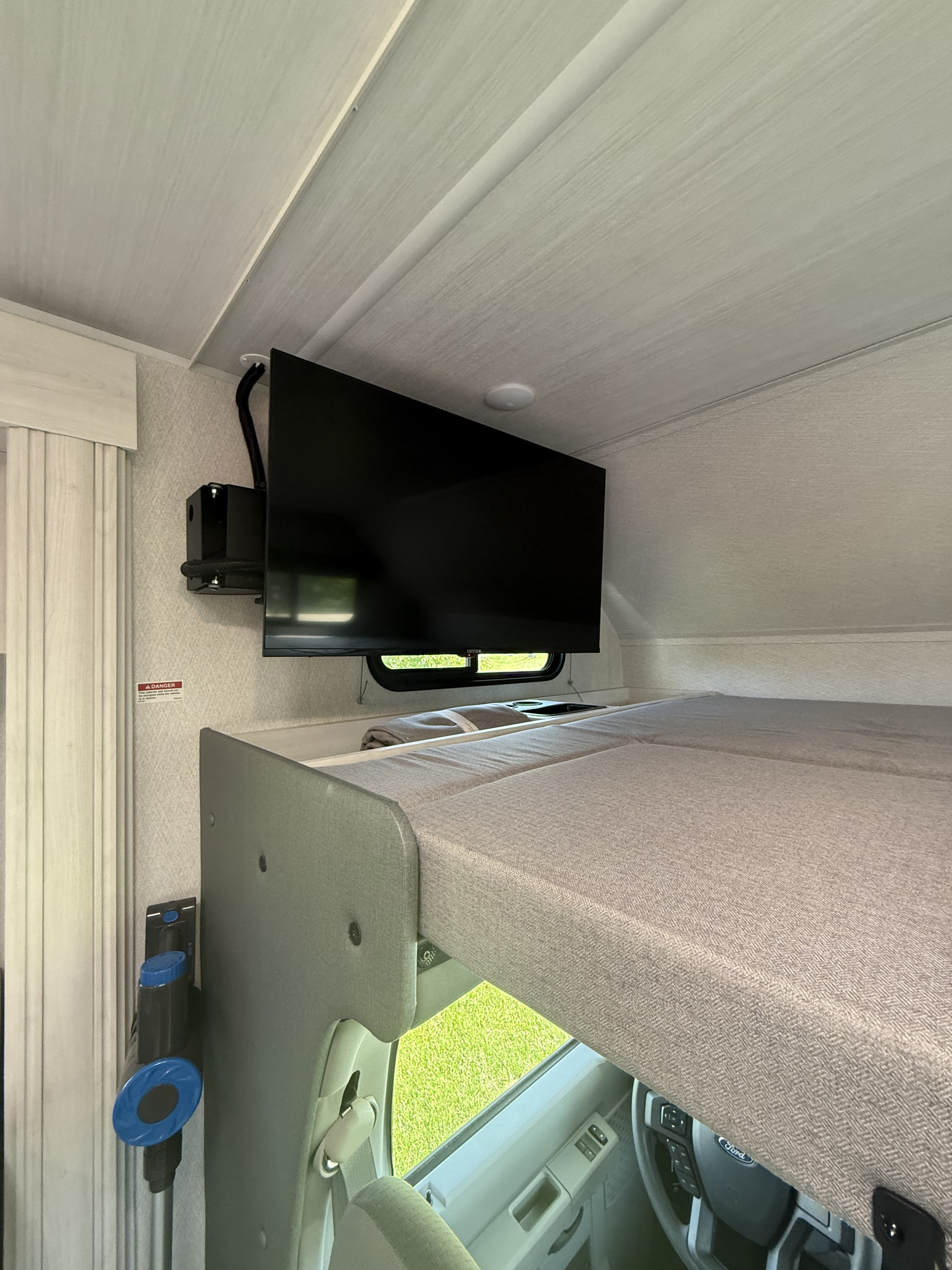 Cozy RV rental interior: overhead bunk bed with cushions, mounted TV, and view into the driver's cab.. Forest River East to West Entrada 2024