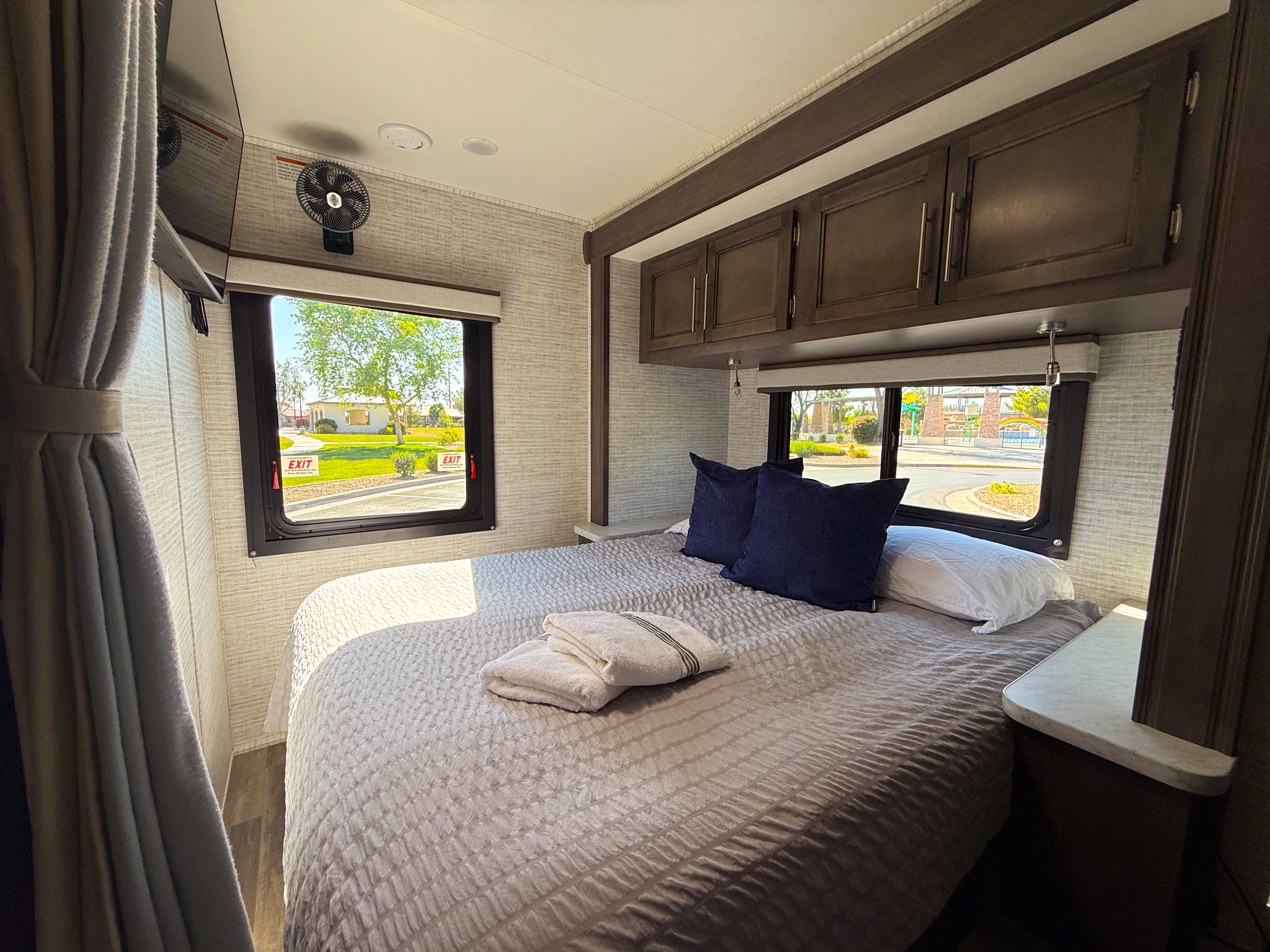 Inside a bright RV rental bedroom: a bed with plush bedding, overhead storage, and large windows revealing a scenic outdoor view.. Entegra Odyssey SE 2025