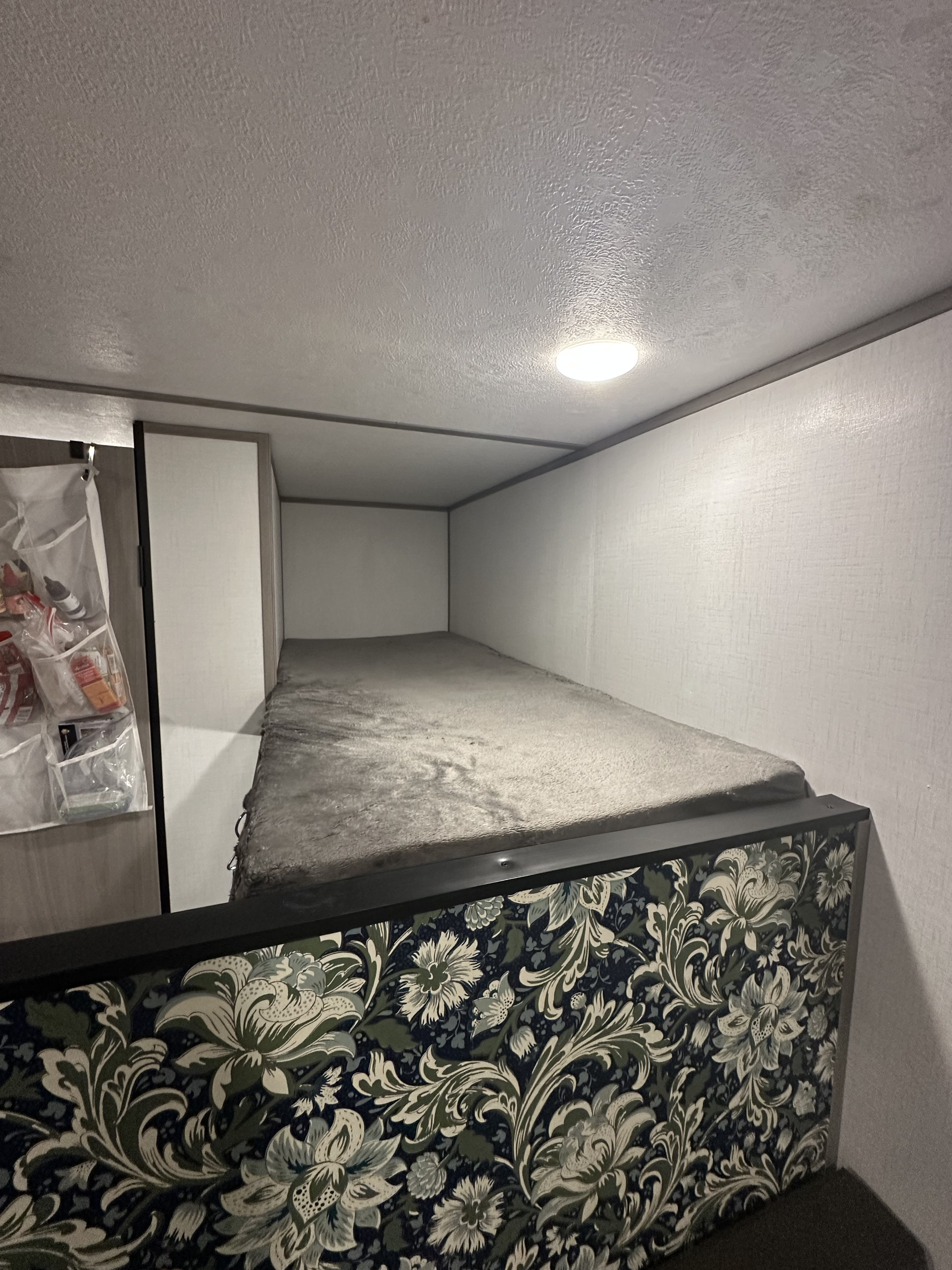 RV rental bunk bed features a floral lower panel, soft grey bedding, textured walls, and a ceiling light. Practical and cozy.. Coleman Lantern 17B 2024