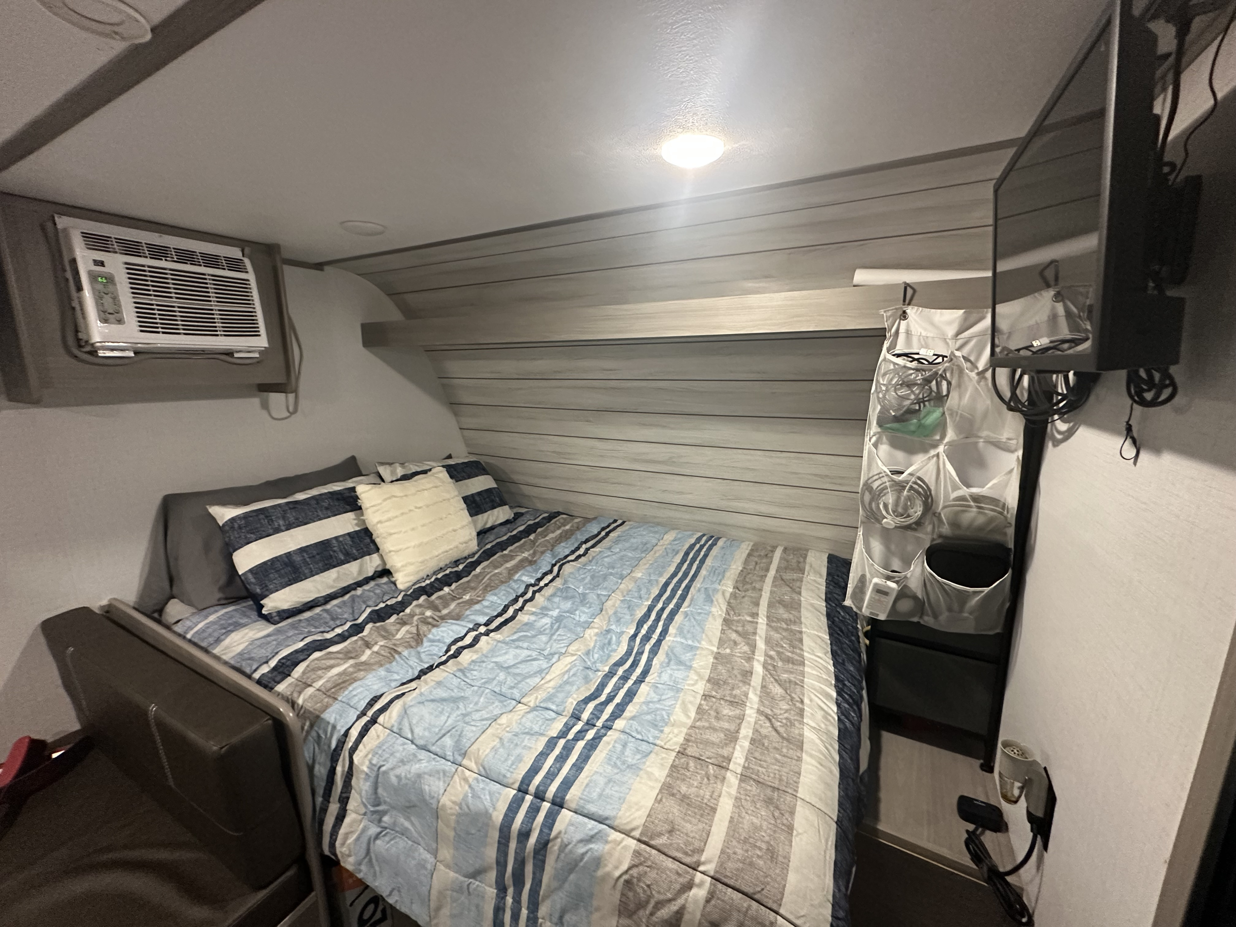 Cozy RV rental bedroom with striped bed, wall-mounted AC, TV, and handy storage, ideal for compact living on the road.. Coleman Lantern 17B 2024