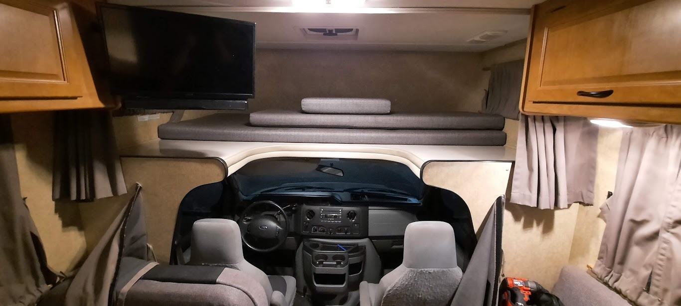 Inside an RV rental: Cozy cab-over bunk with stacked cushions and a TV, overlooking the vehicle's dashboard and steering wheel.. Thor Majestic 2017