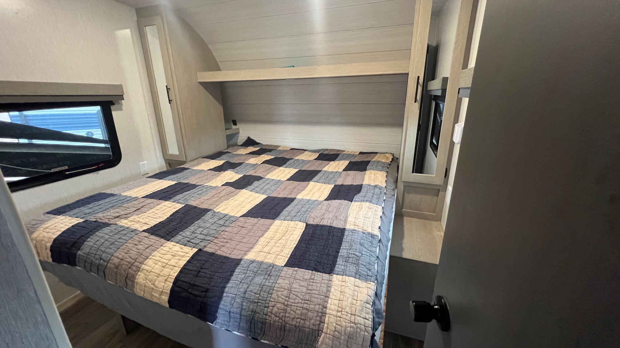 Cozy RV rental bedroom with a checkered quilt, overhead storage, and window. Compact yet comfortable interior for travel.. Forest River Stealth 2023