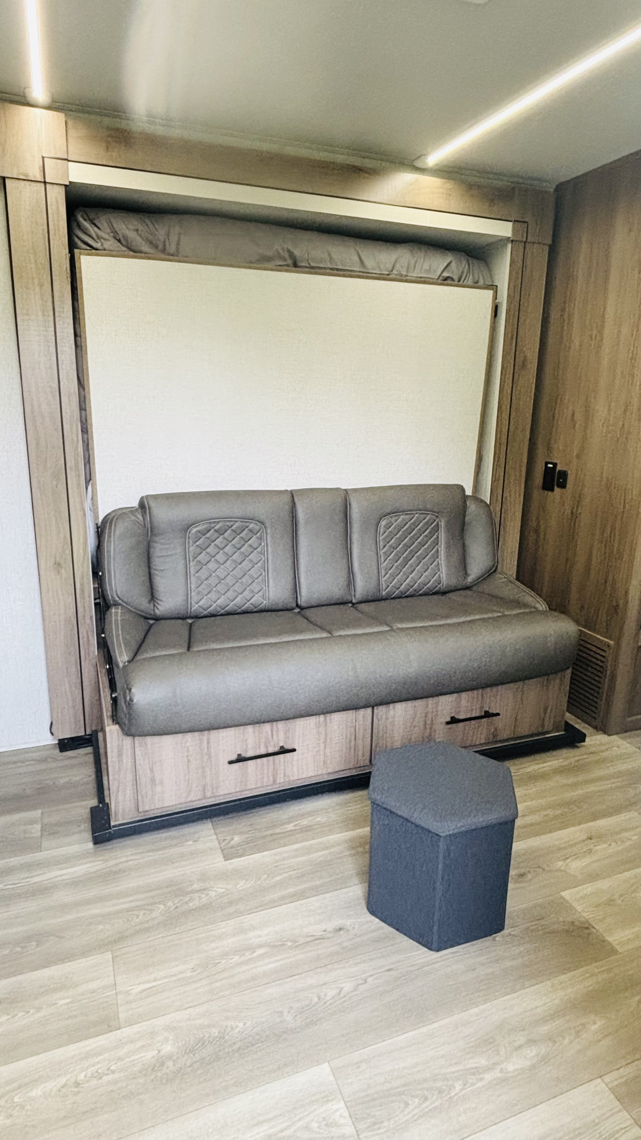 Modern RV rental interior with a gray sofa, pull-down murphy bed, and storage drawers. Hexagonal ottoman on wood floor.. Grand Design Imagine AIM 2024