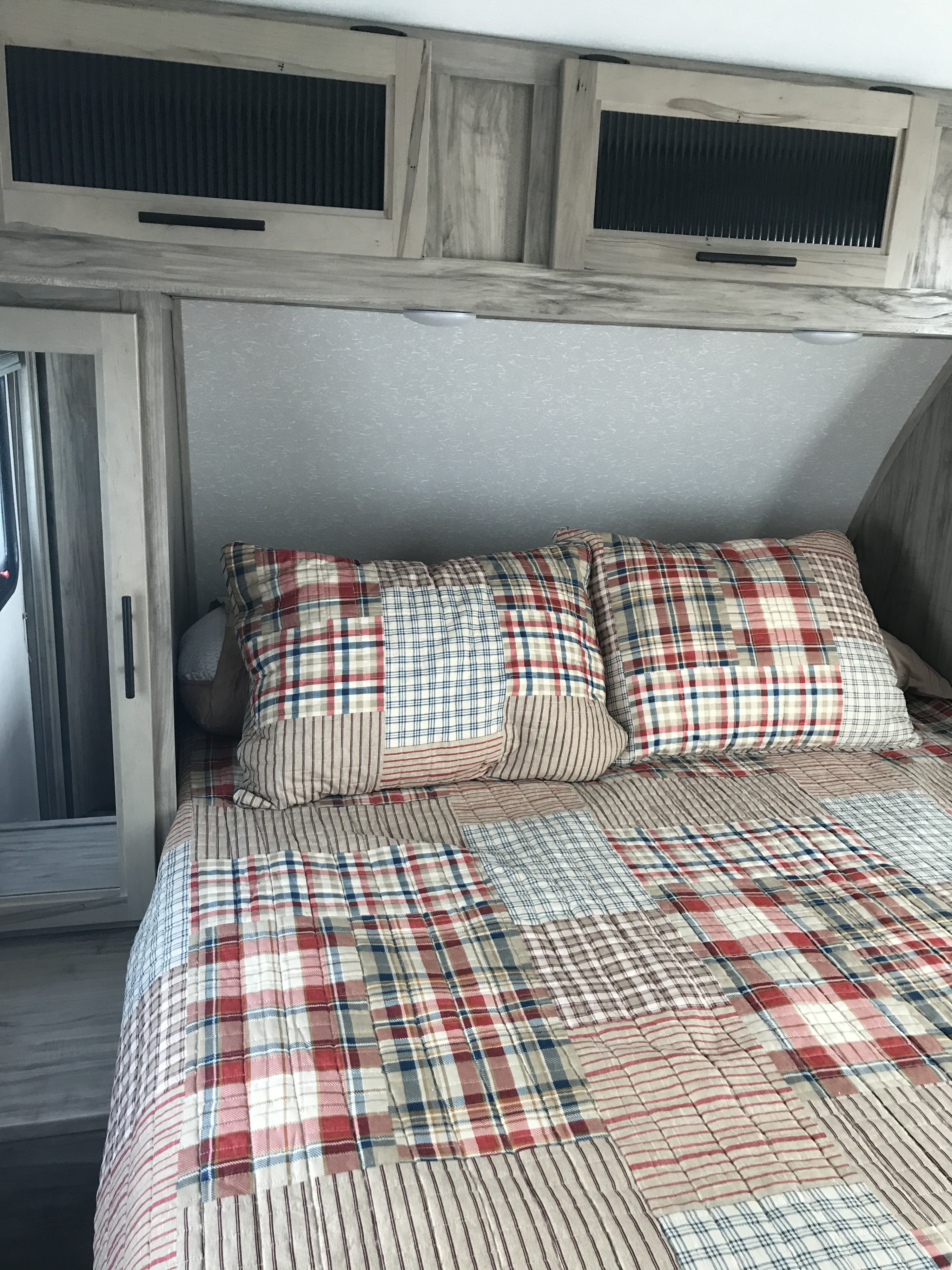 Inviting RV rental bedroom with a cozy patchwork plaid quilt, matching pillows, and modern light wood overhead cabinets.. Forest River Cherokee 2021