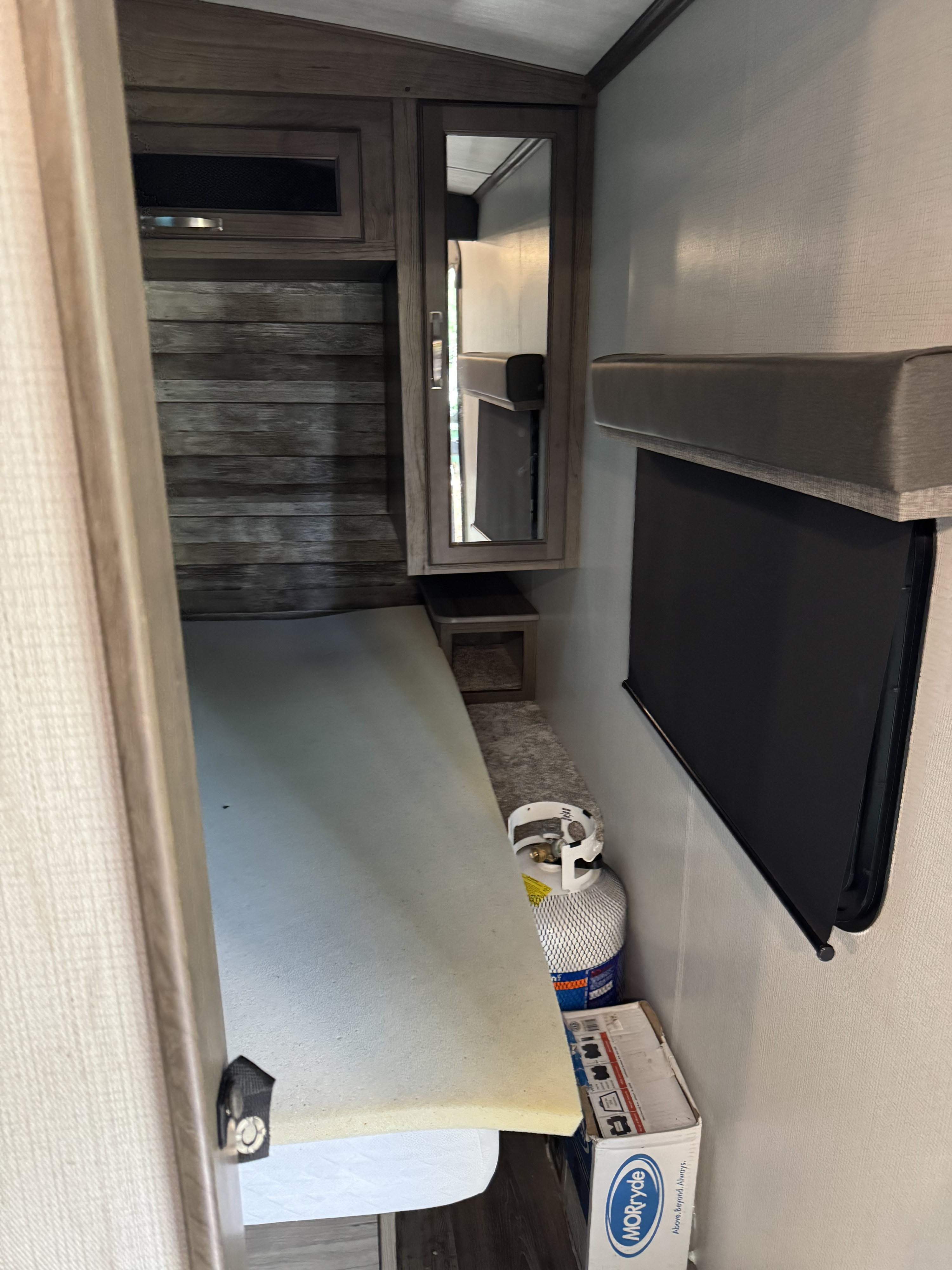 This RV rental features a cozy bedroom with a bed, wood-paneled wall, mirrored cabinet, and a black window blind. Storage & tank visible.. Keystone RV Cougar 2022