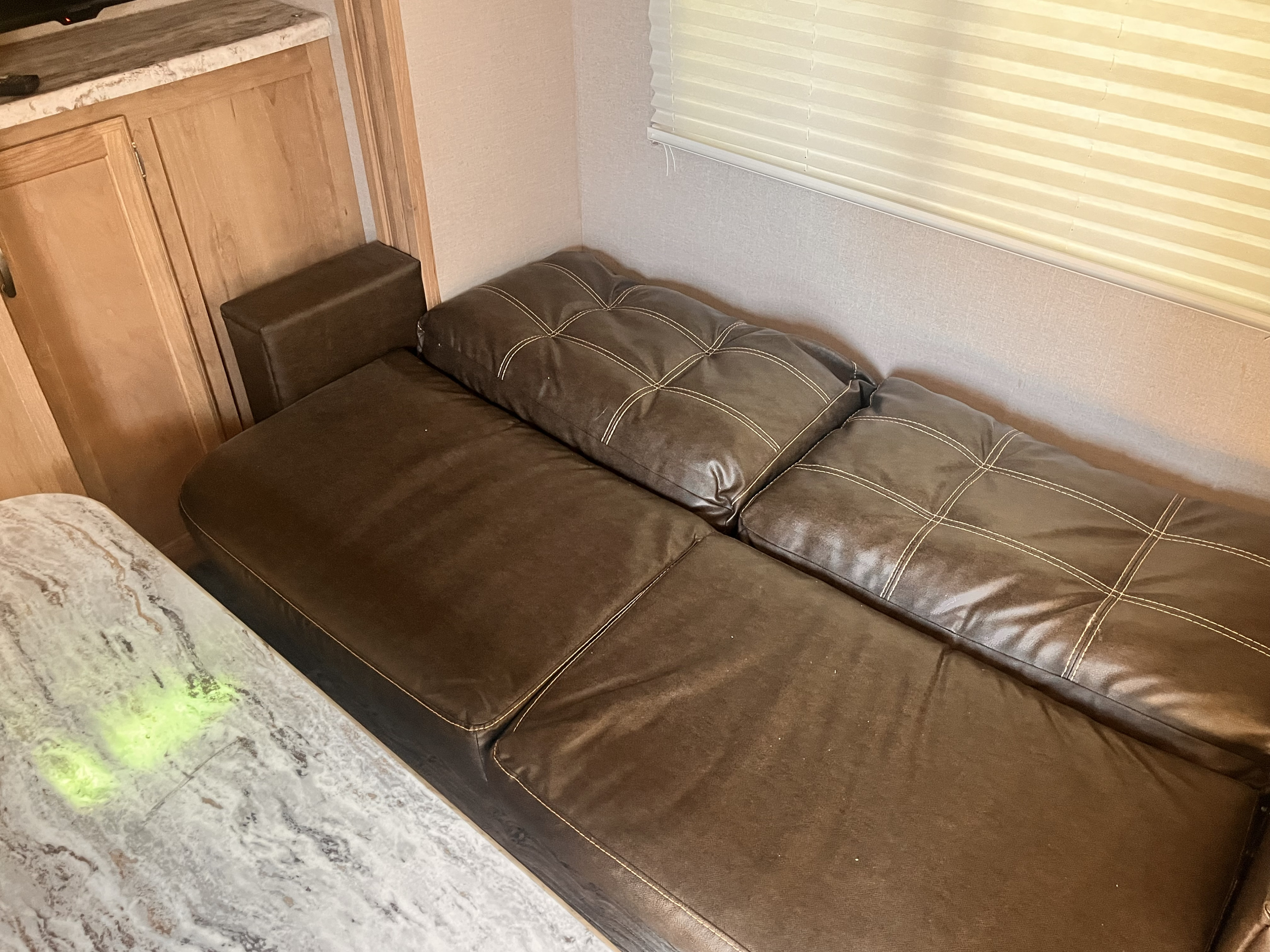 RV rental interior: A comfy brown leatherette sofa with quilted cushions, near wooden cabinets, and a window with blinds.. Minnie Micro Minnie 2020