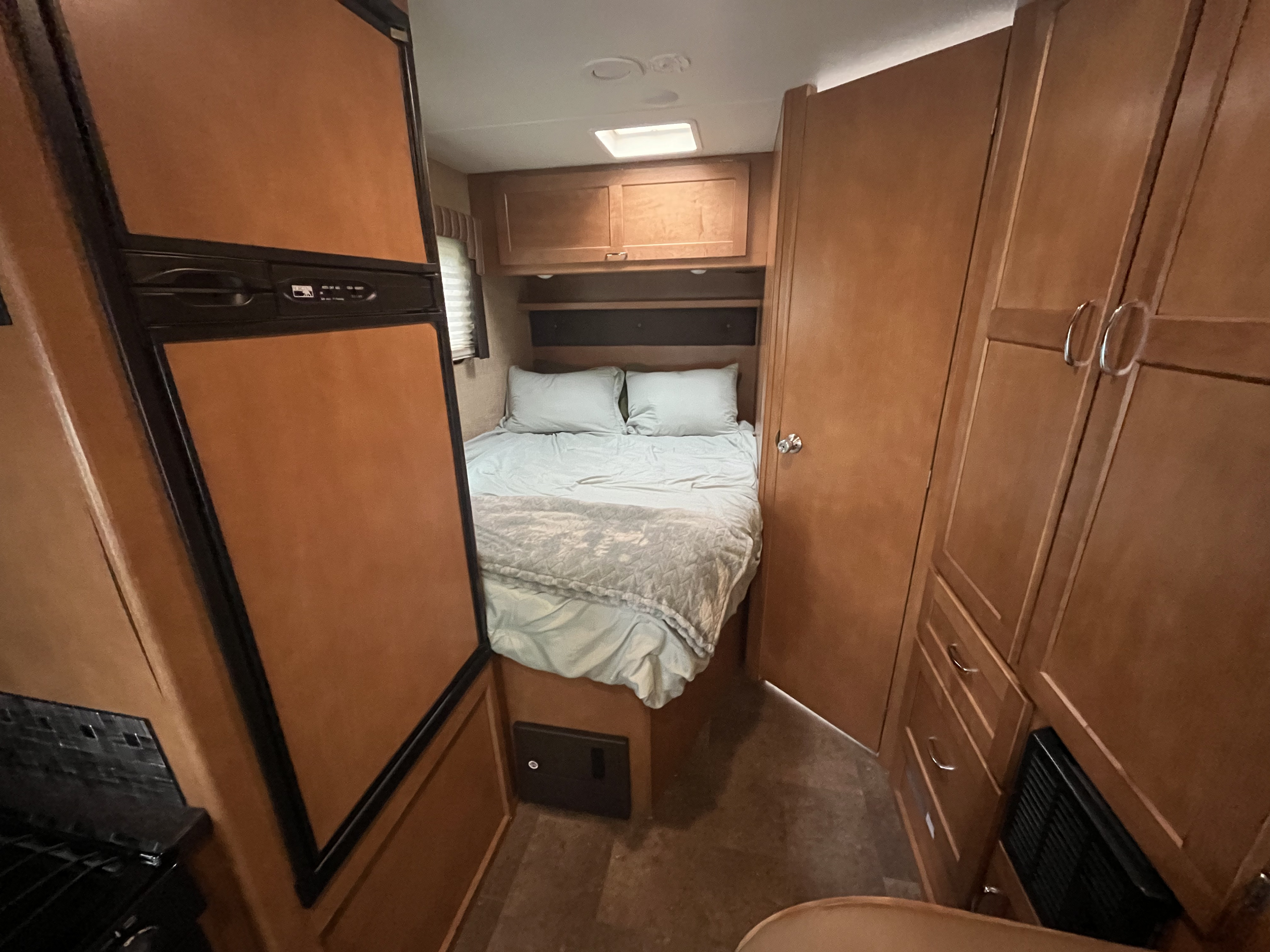 Interior of an RV rental showing a compact bedroom with a bed and overhead cabinets. A refrigerator and additional storage are visible.. Winnebago Minnie Winnie 2018
