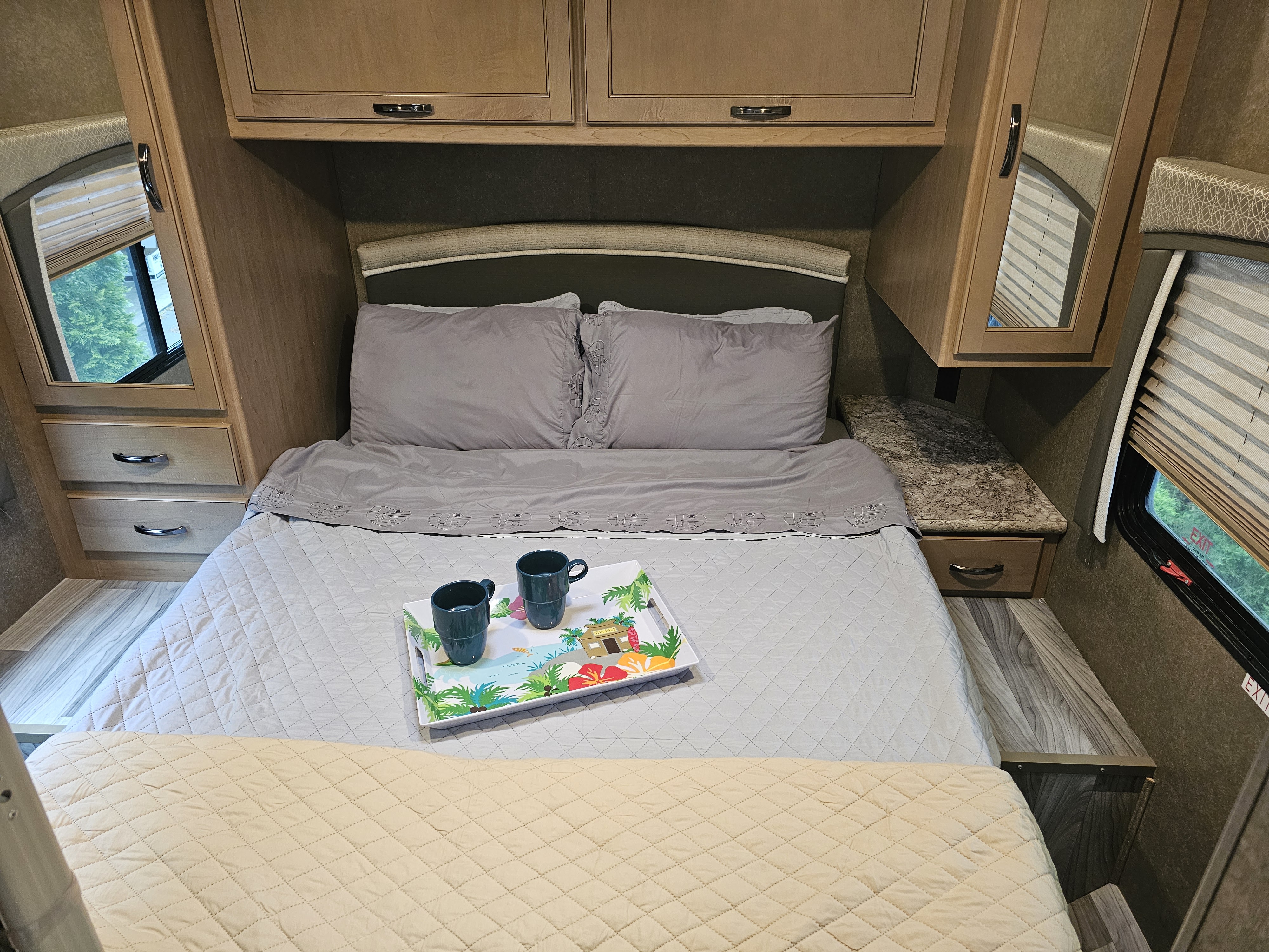 Cozy RV rental bedroom with a gray-sheeted bed, tropical tray, overhead cabinets, and side storage units with mirrors.. Thor Four Winds 2021