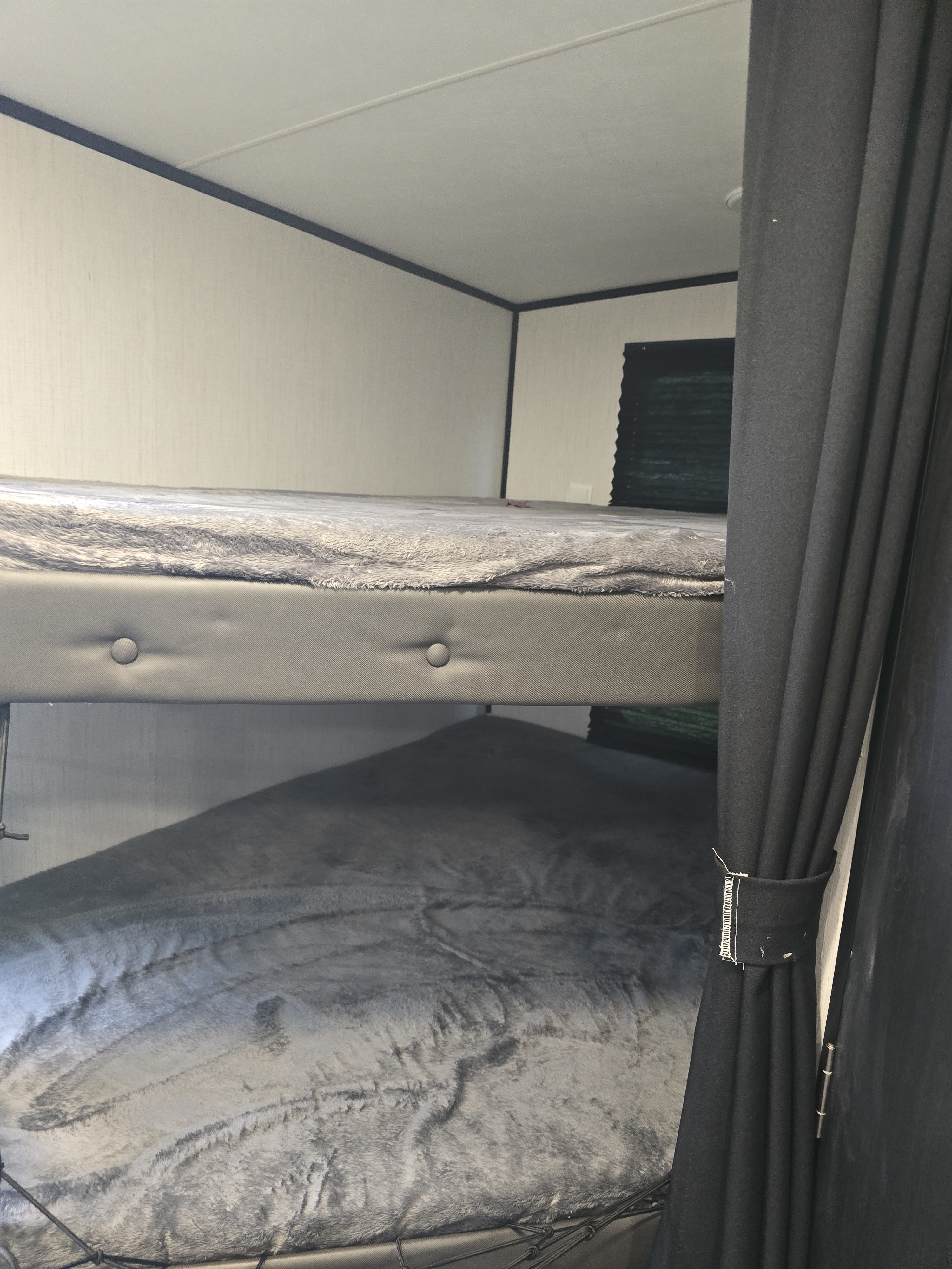 Interior view of an RV rental showing stacked bunk beds with soft grey blankets, dark privacy curtains, and light walls.. Heartland RVs Mallard 2022