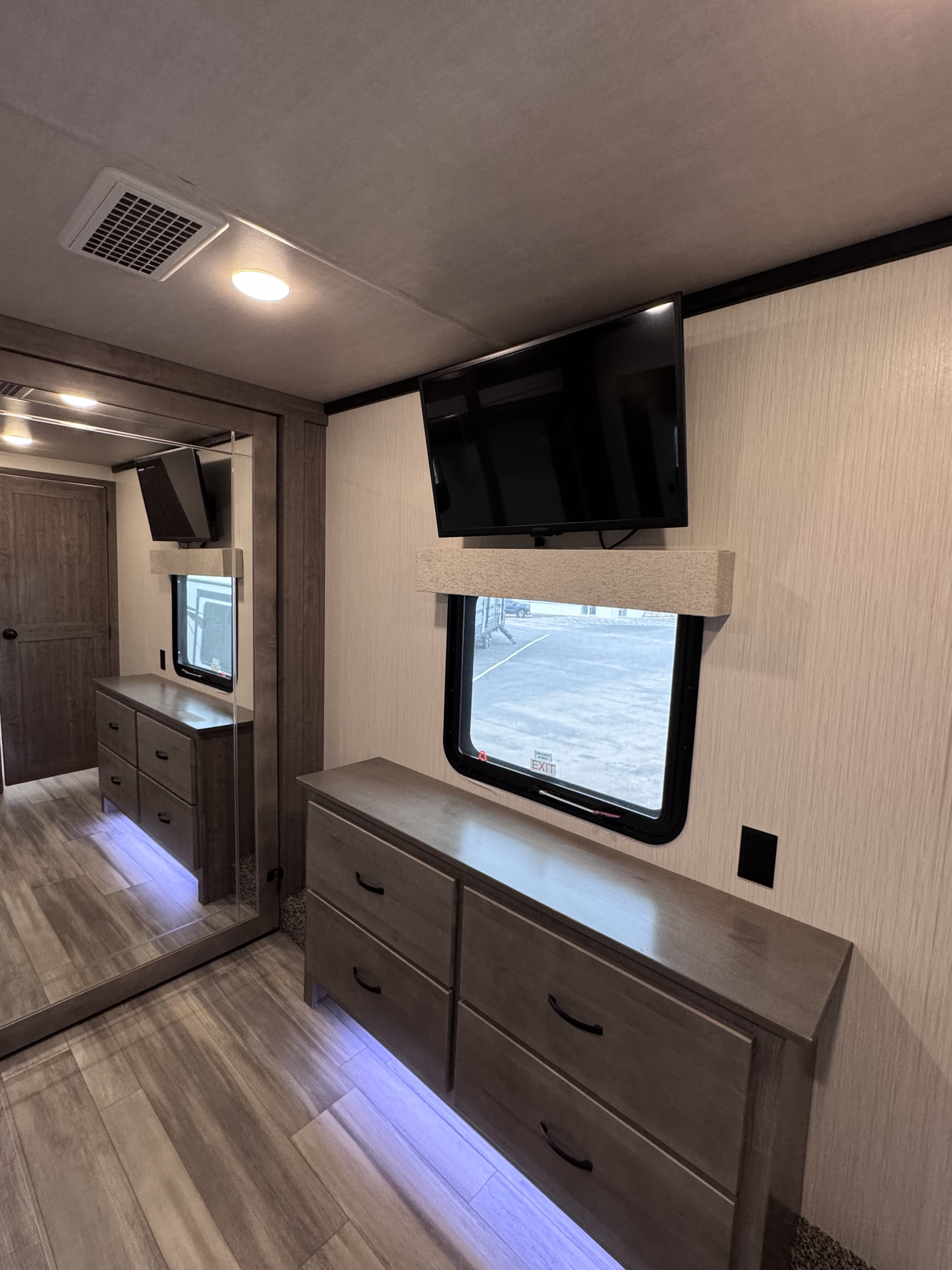 RV rental bedroom with wall-mounted TV, long dresser with accent lights, window, and large mirrored closet.. Forest River Cedar Creek 2022