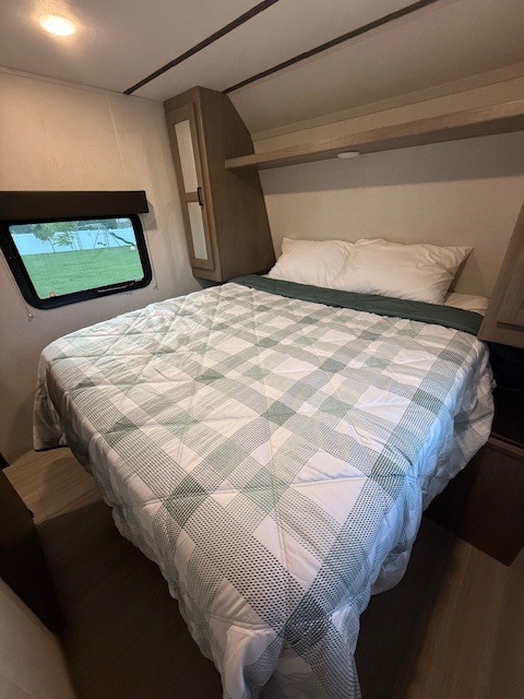 Comfortable RV rental bedroom featuring a plaid bed, overhead storage, and a window with a scenic outdoor view.. Keystone RV Hideout 2022