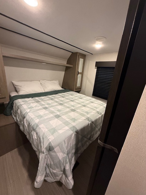 Interior view of an RV rental bedroom, featuring a cozy bed with a plaid comforter, overhead storage, and a window.. Keystone RV Hideout 2022