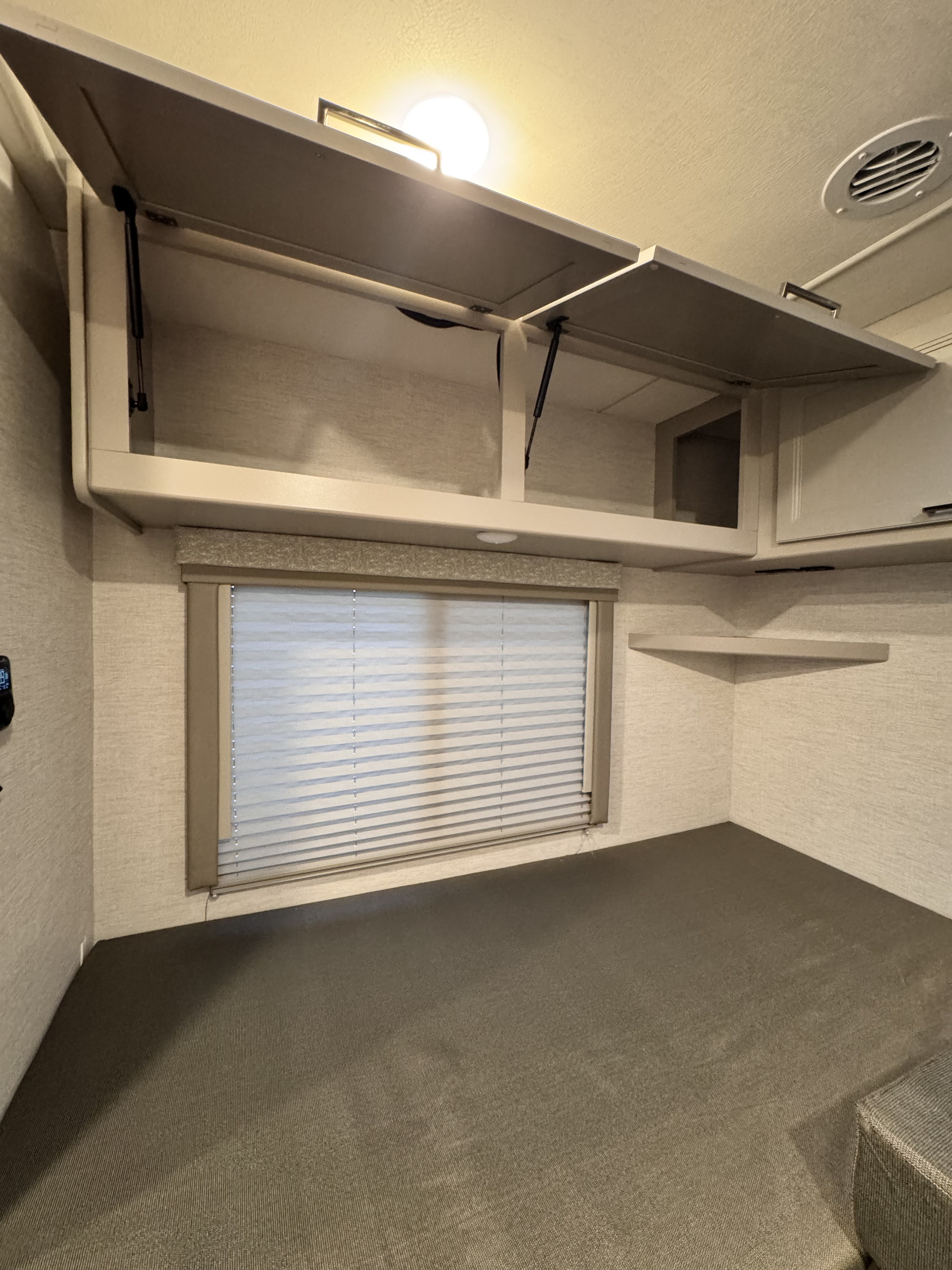 Inside an RV rental: a compact sleeping area with open overhead storage, a window with blinds, and a corner shelf.. Thor Motorcoach Four Winds 2025
