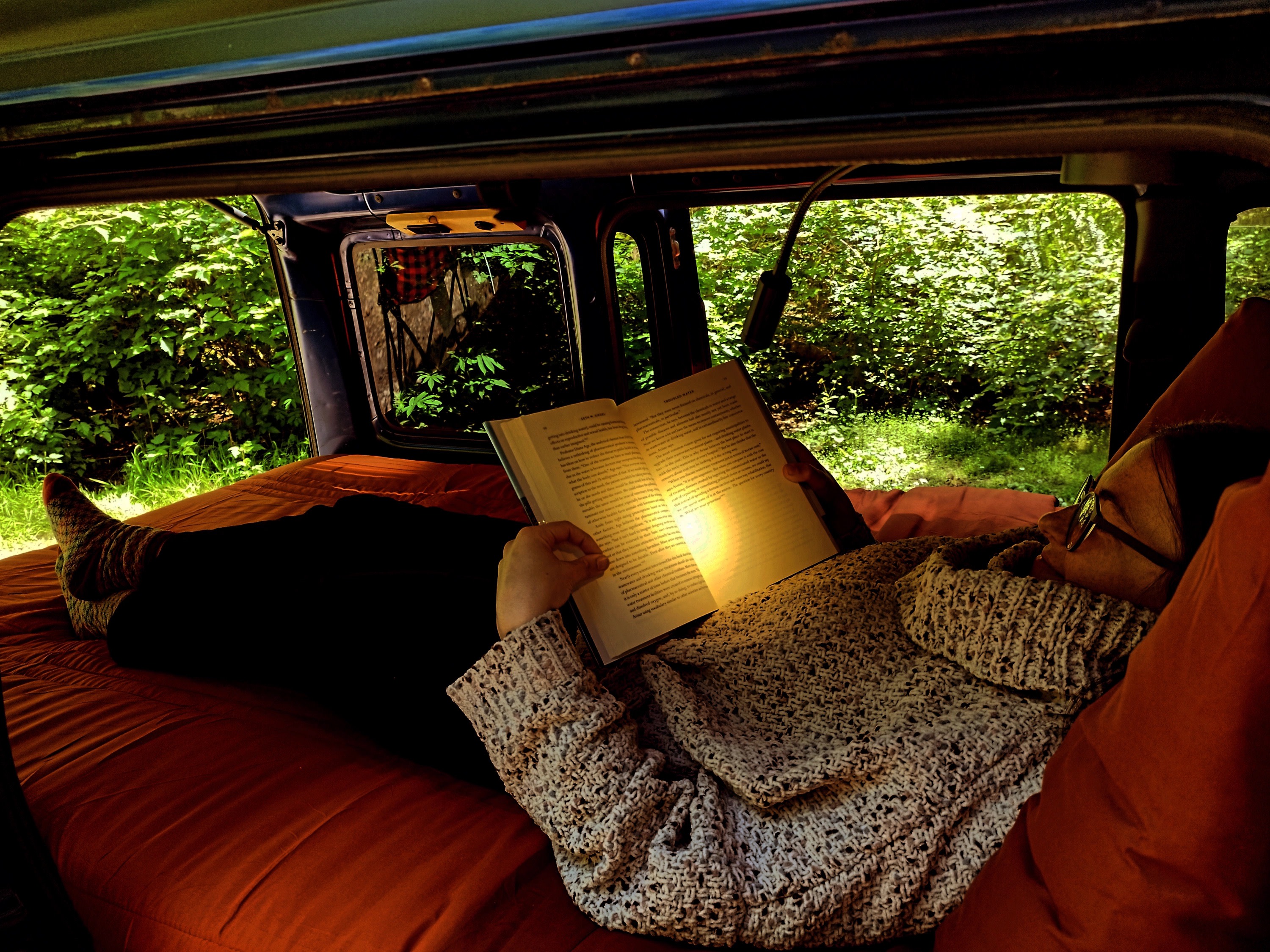 Inside an RV rental, a person lies on an orange bed, reading by a lamp, with lush green nature visible outside.. Daihatsu Hijet Cargo 2007