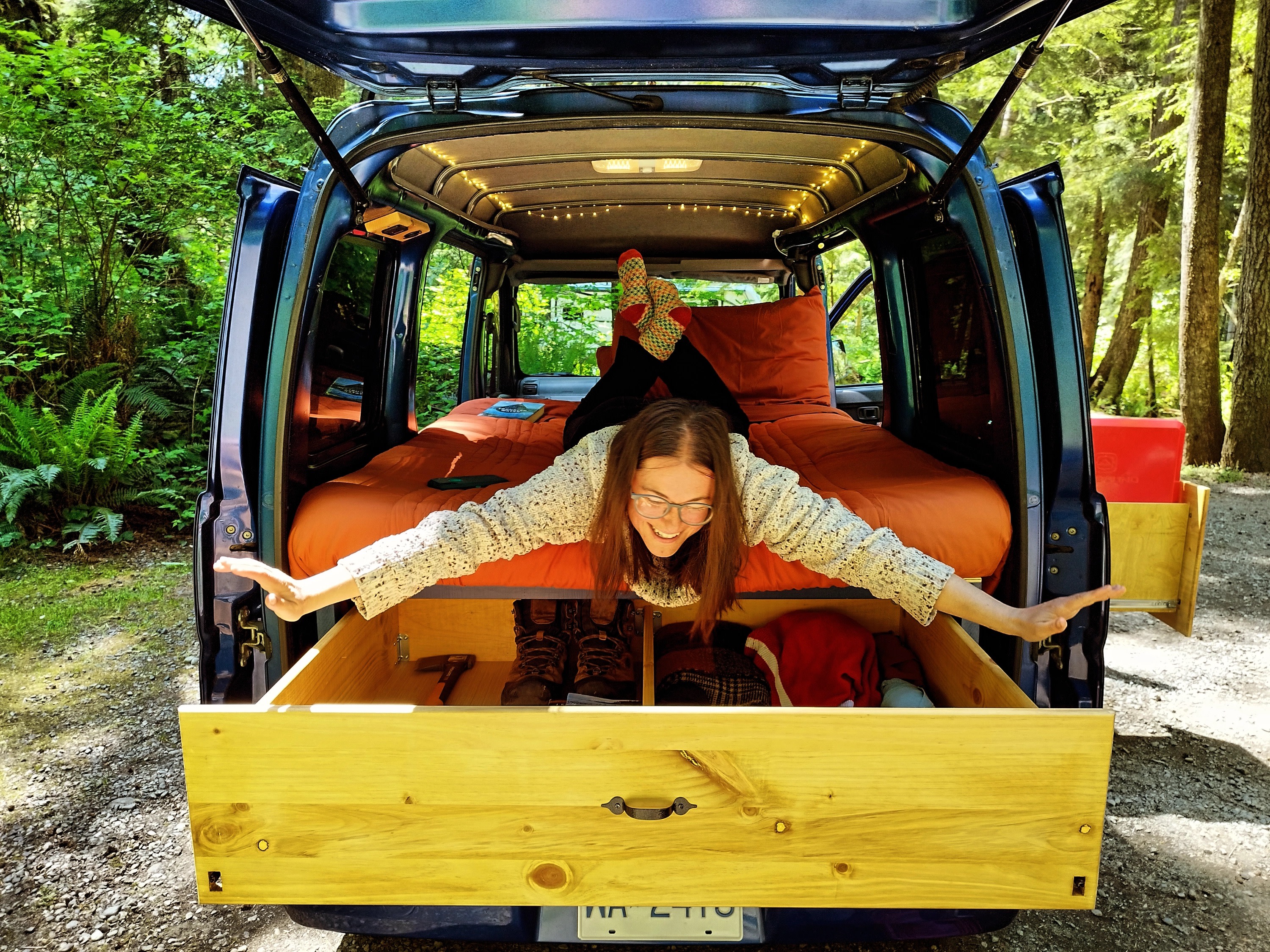 Experience an RV rental! A smiling woman stretches out in her camper van, featuring a cozy bed & convenient pull-out storage.. Daihatsu Hijet Cargo 2007