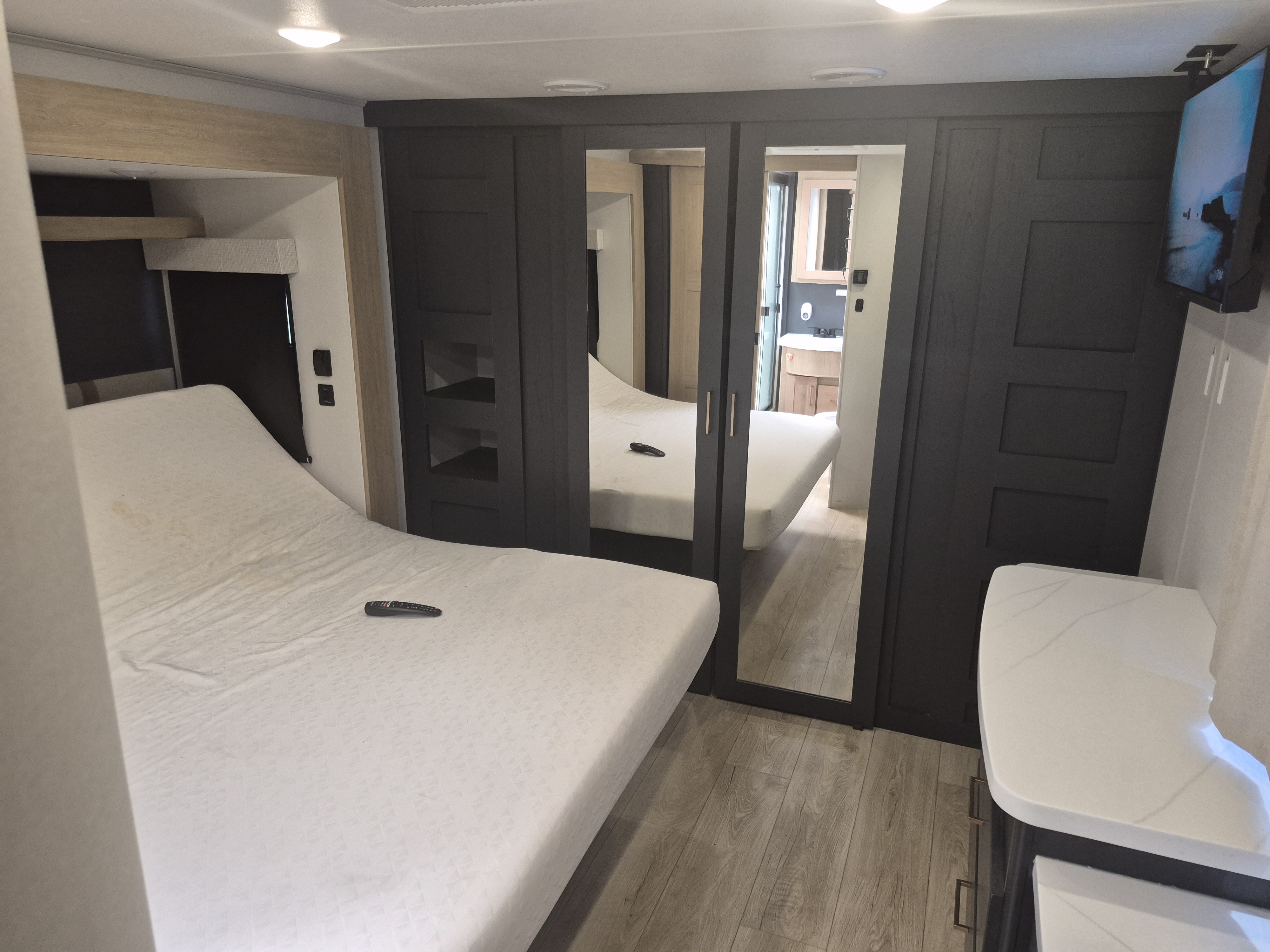 Modern RV rental bedroom with a comfy bed, dark wood cabinets with mirrors reflecting the bed & bathroom, plus a wall TV.. Heartland RVs Cyclone Toy Hauler 2024