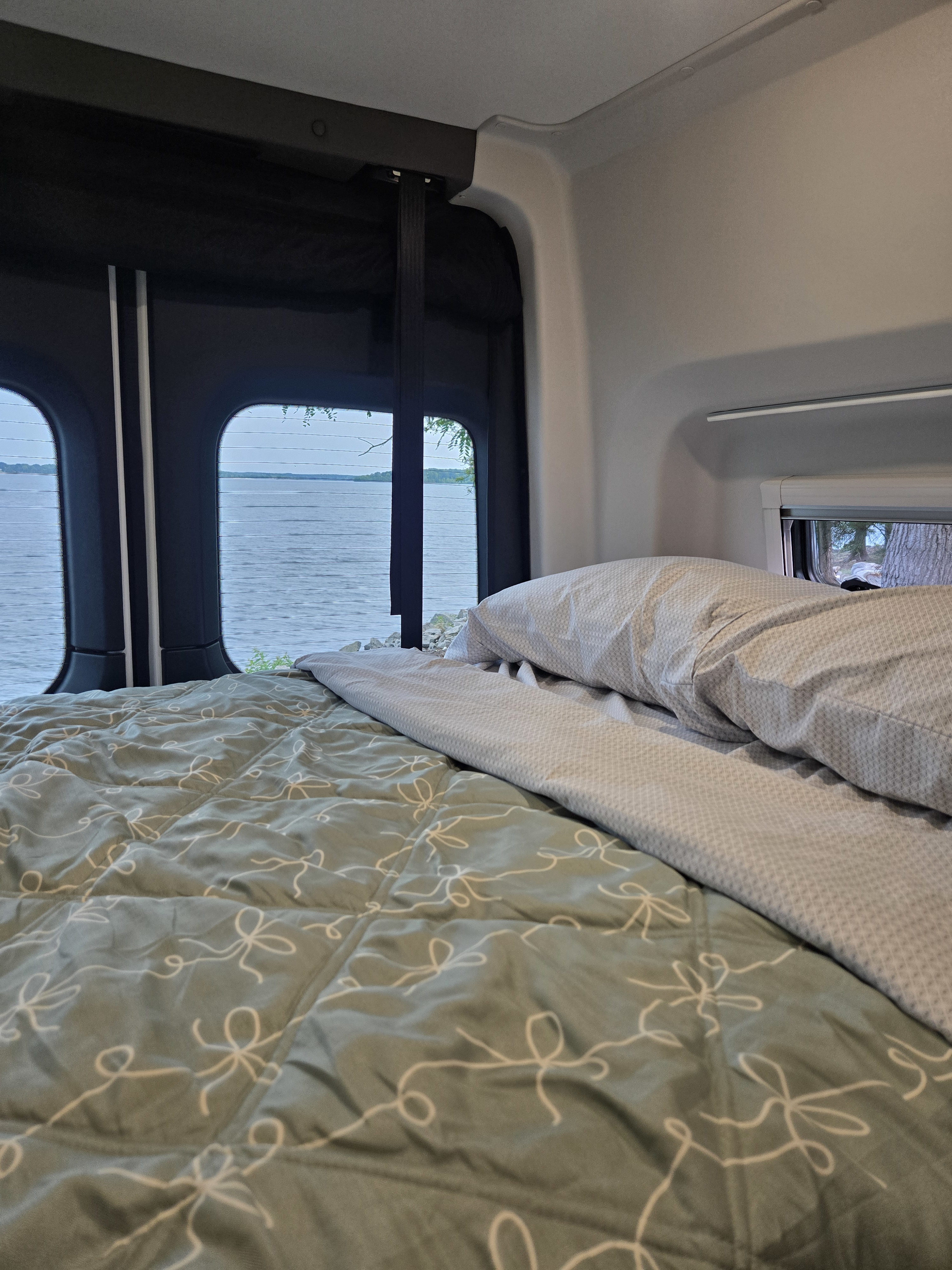 RV rental: A cozy bed with a green comforter and patterned pillow overlooks a calm lake through wide rear windows.. GH1 GH1 Class B 2025
