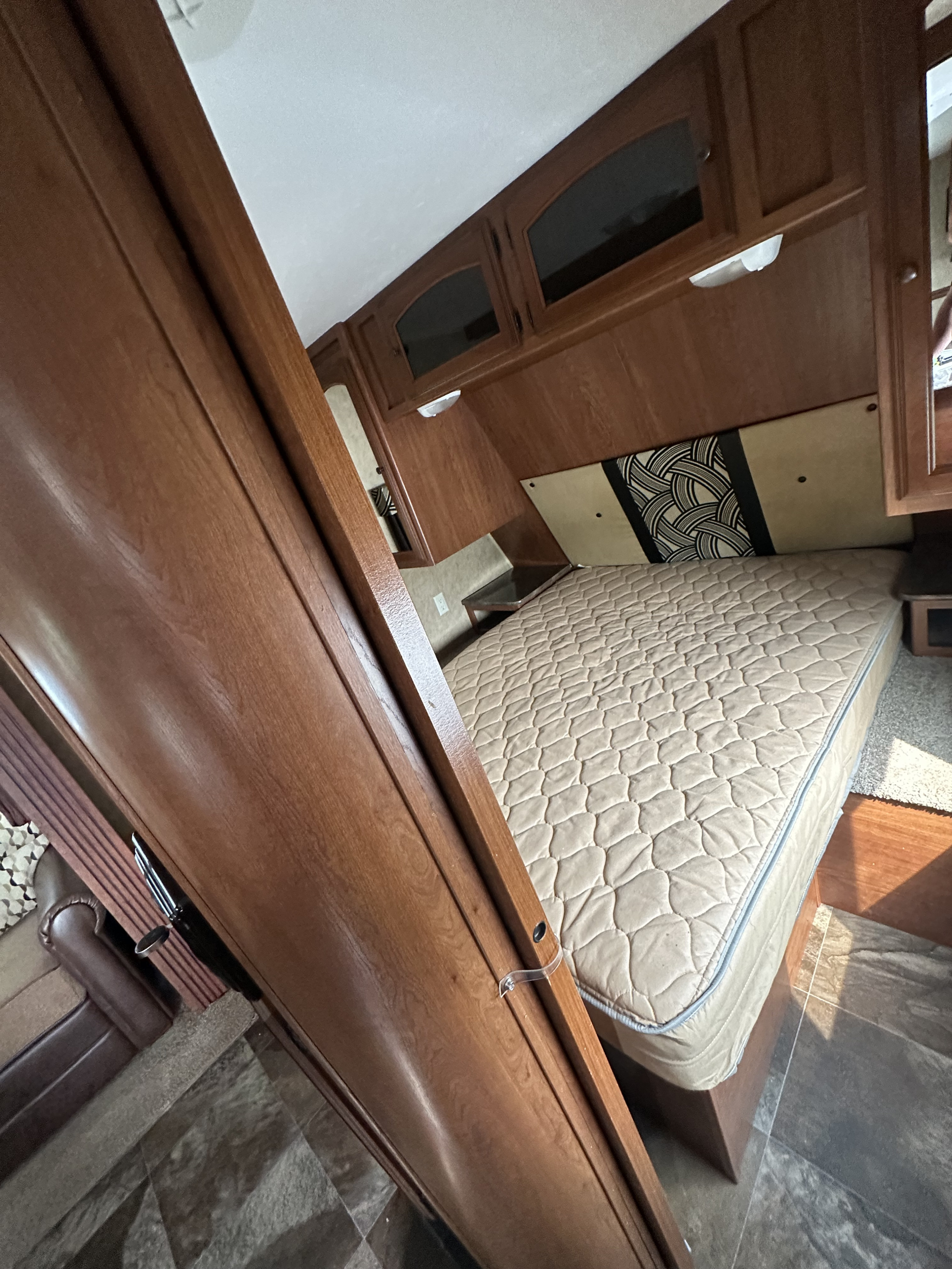 Cozy RV rental bedroom featuring a comfortable mattress, patterned headboard, and overhead wood cabinets. Ideal compact living space.. Dutchmen Classic 2013