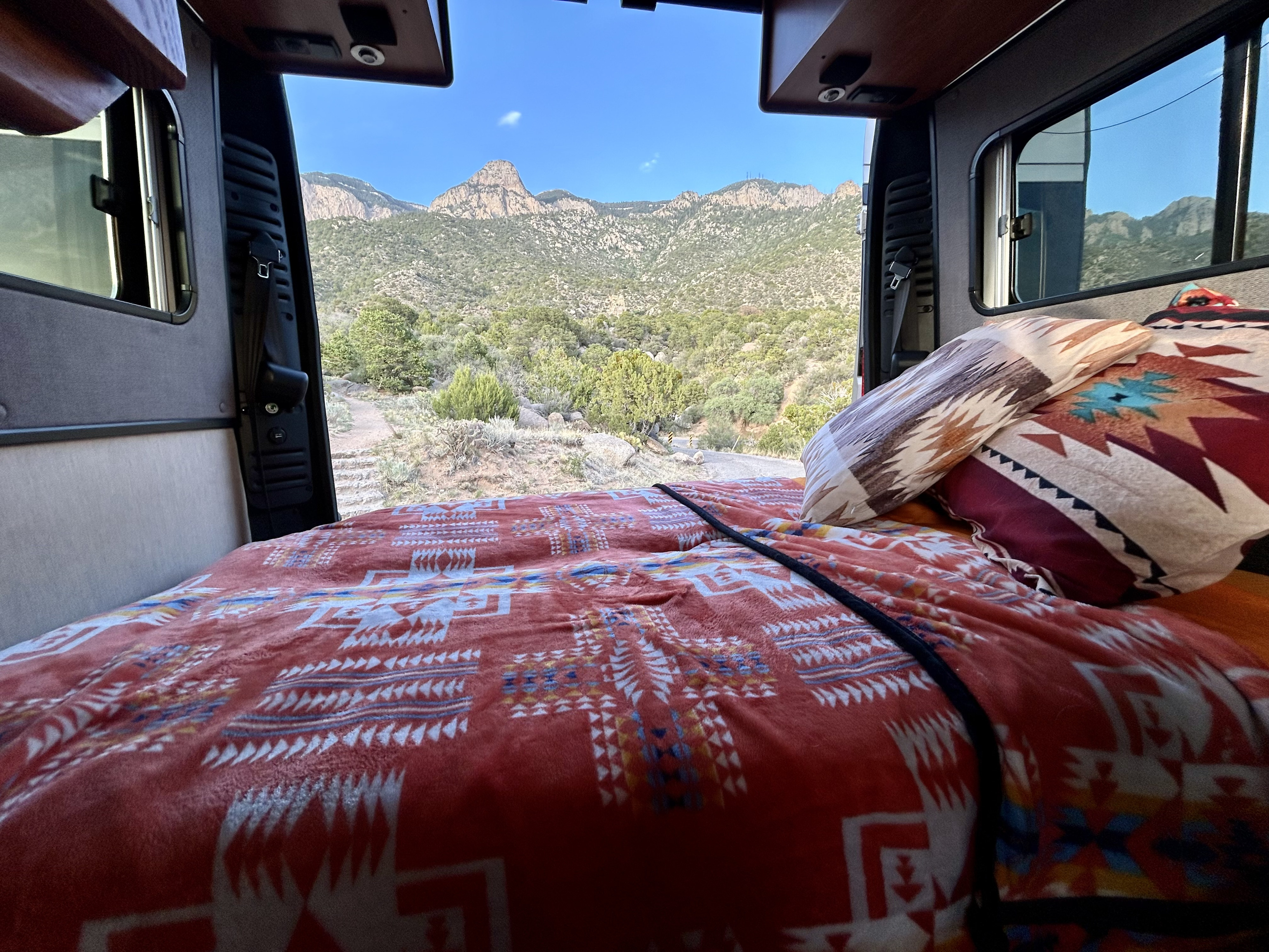 Inside a comfy rv rental, a patterned bed faces open doors revealing stunning mountains and a blue sky.. Roadtrek Roadtrek Class B 2019