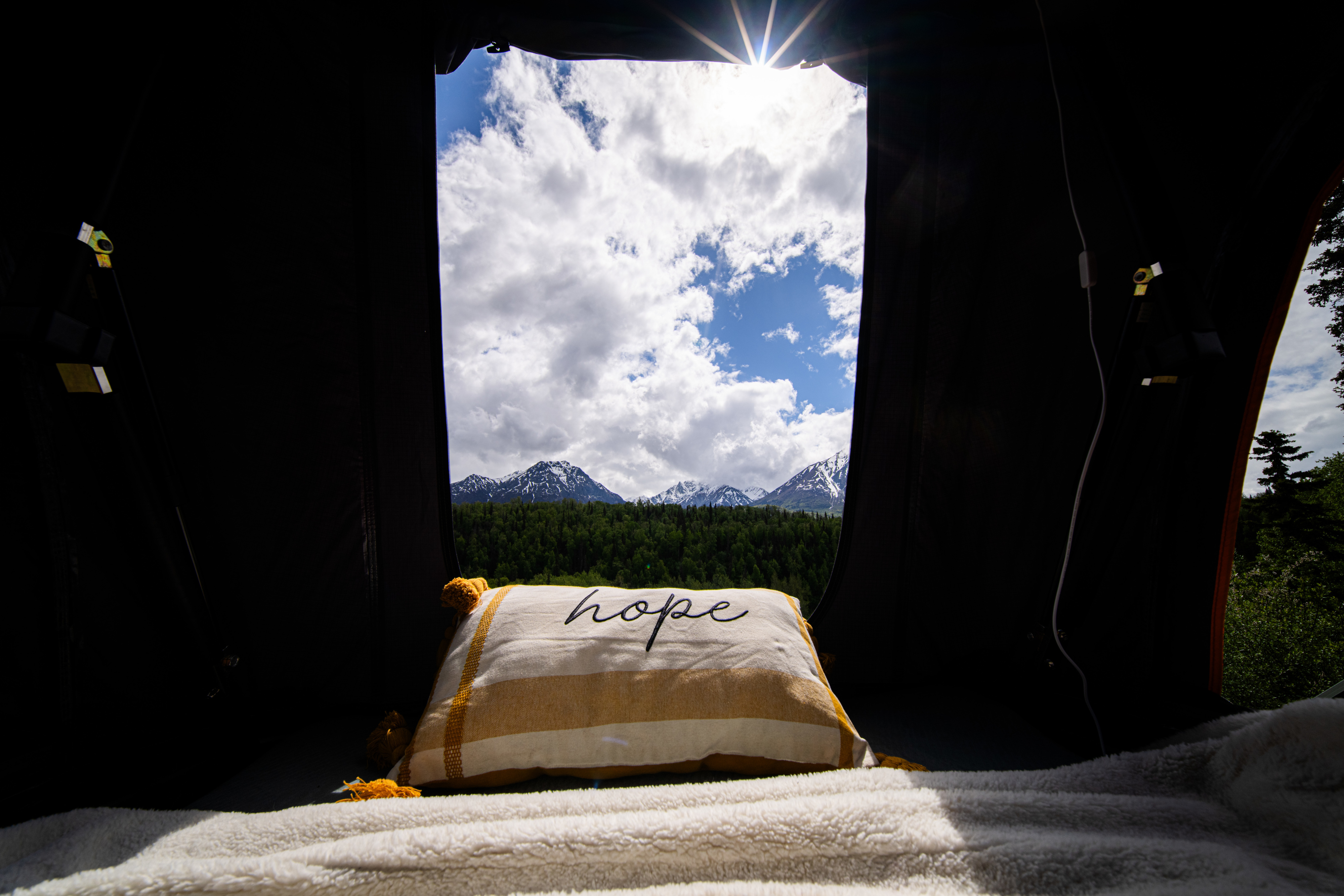View from an RV rental: A 'hope' pillow on a cozy bed looks out to snow-capped mountains, green forest, and bright, cloudy sky.. Honda Element 2008