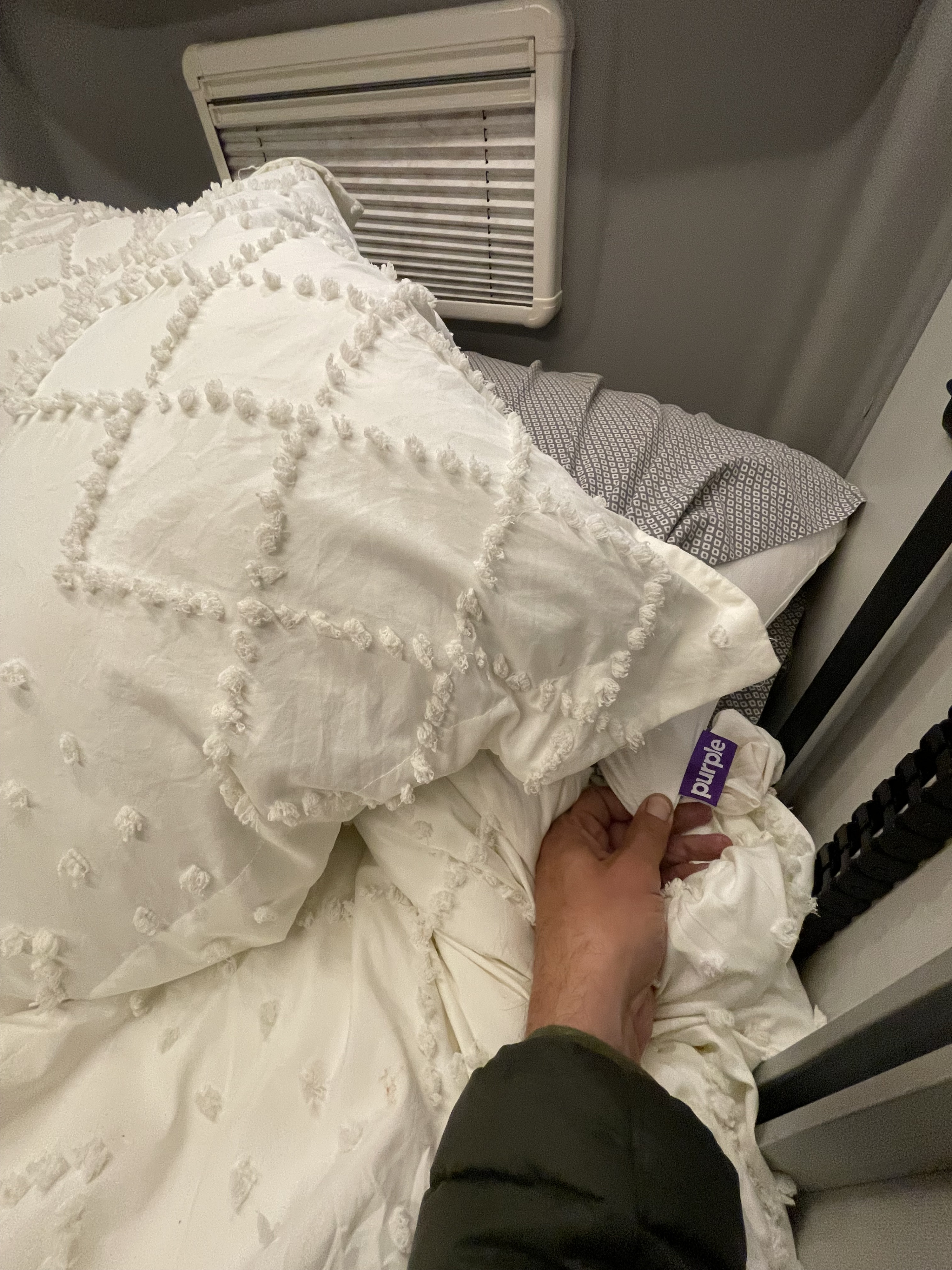 Hand holding Purple brand tag on textured white bedding in an RV rental. Window blinds & grey patterned pillowcase visible.. Winnebago Revel 2019
