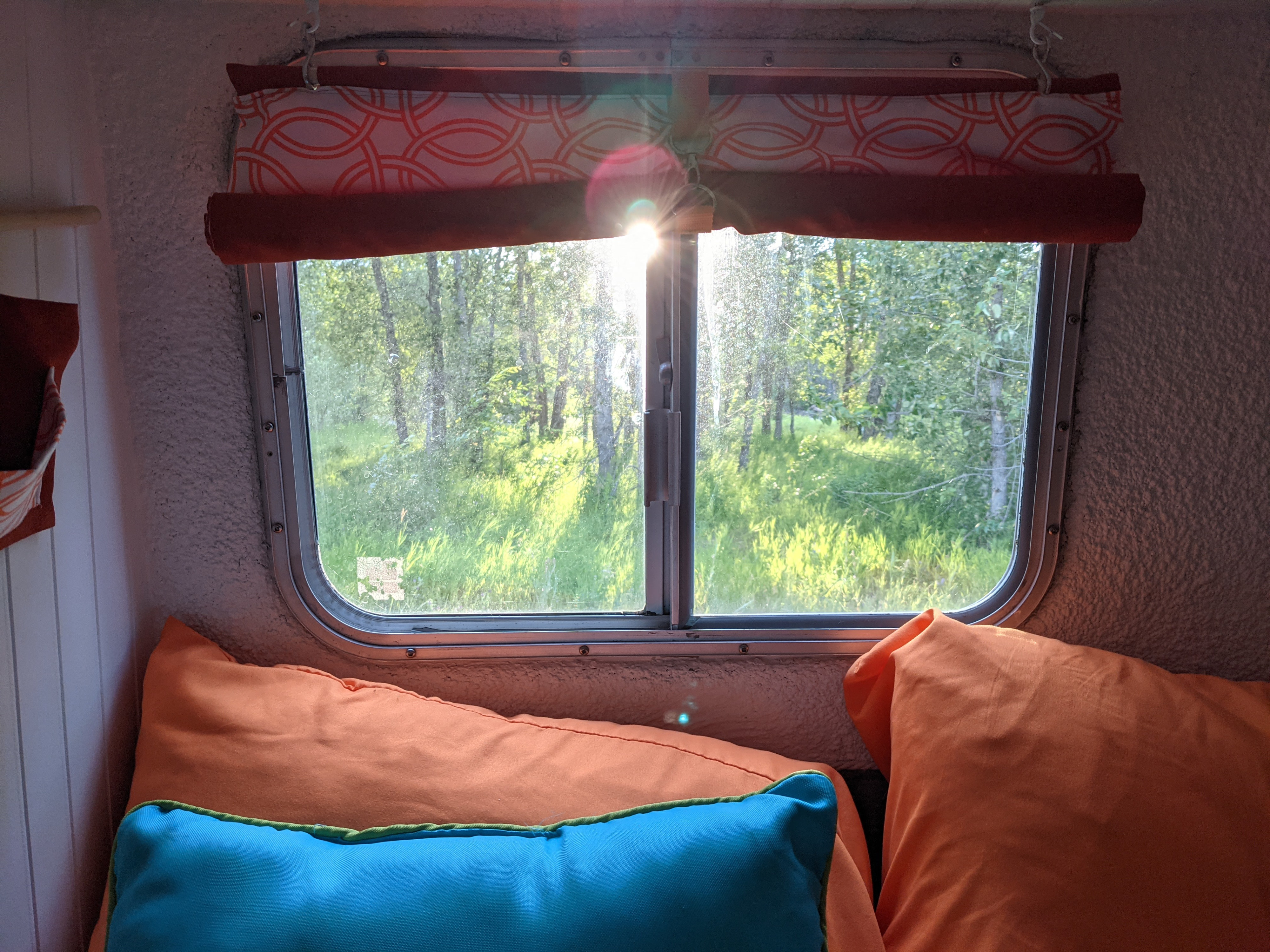 Inside an RV rental: bright sun through the window illuminates lush green nature. Orange and blue pillows add comfort.. Boler Neonex 1300 1979