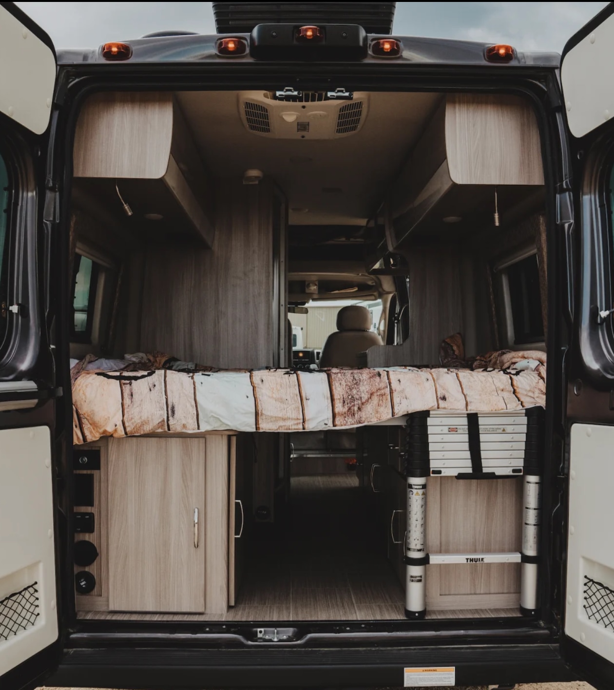 Inside an RV rental: A cozy bed with wood-patterned bedding, clever storage cabinets, and a portable Thule ladder.. Thor Sequence 2022