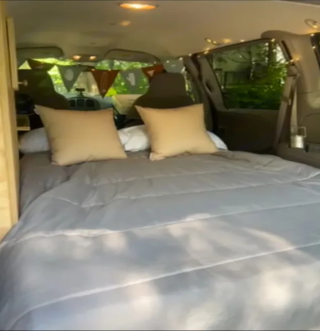 A transformed RV rental van interior showcasing a cozy bed with pillows, decorative pennants, and string lights. Ideal for travel.. Dodge Caravan 1999