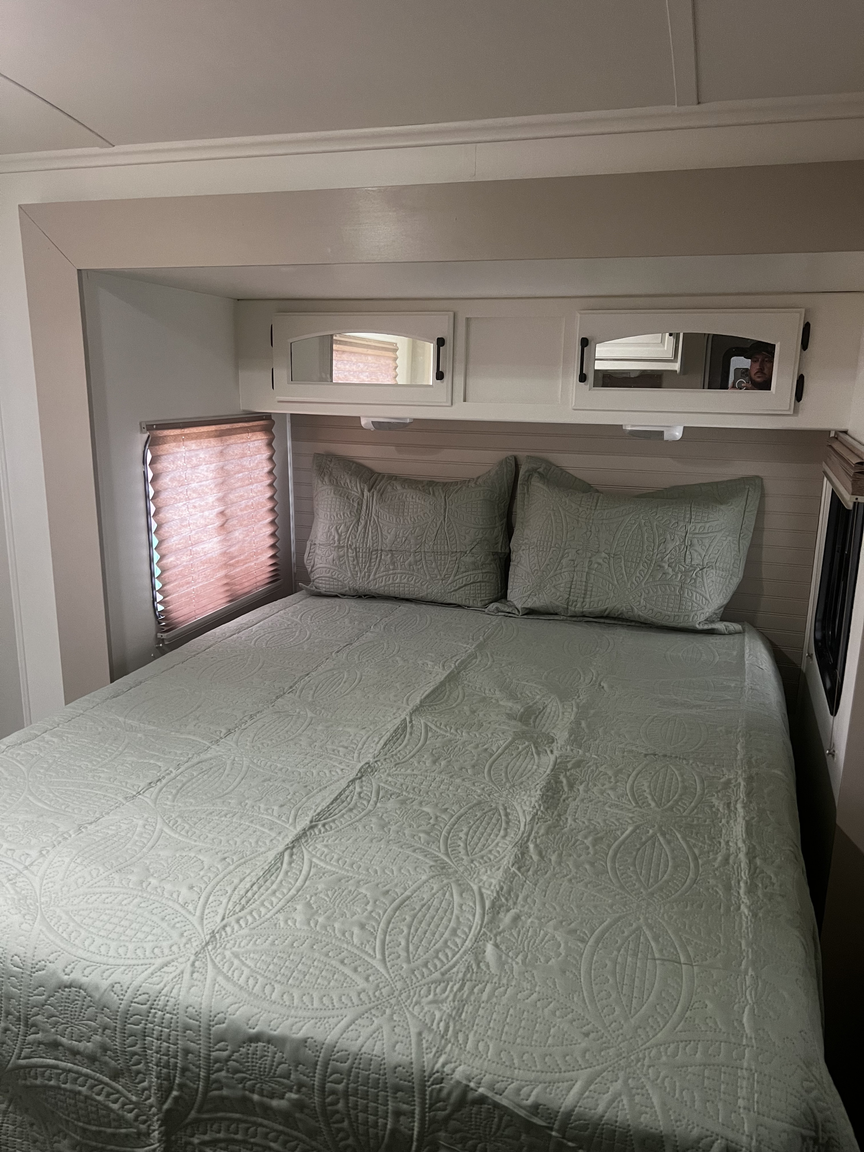 An RV rental bedroom featuring a neatly made bed with light green quilted bedding, two pillows, mirrored cabinets, and a window.. Keystone RV Outback Toy Hauler 2013