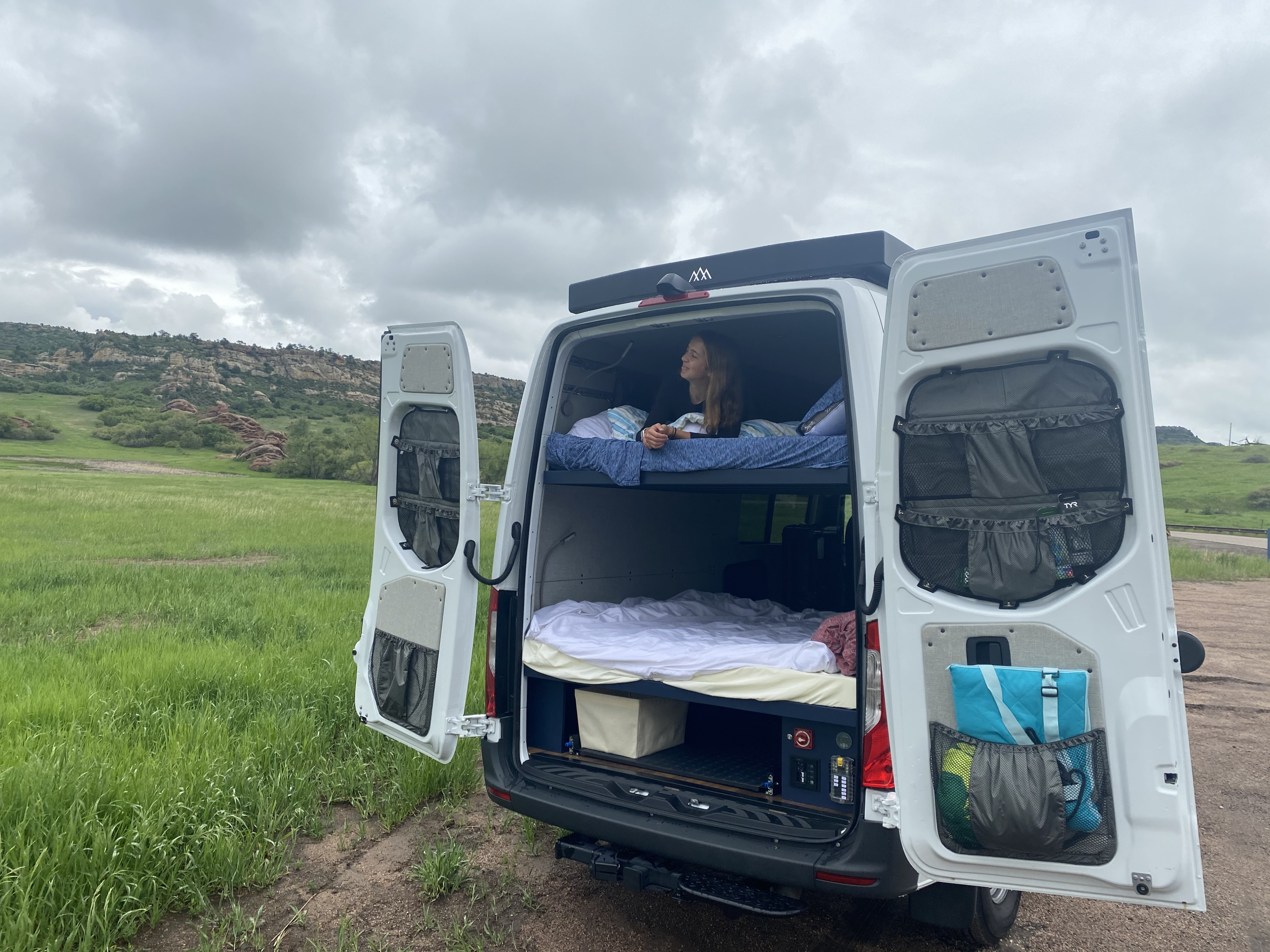 Relaxing in an RV rental van, a woman on the bunk bed enjoys views of a green field and rocky mountains. Perfect for van life.. Mercedes-Benz Sprinter 2021