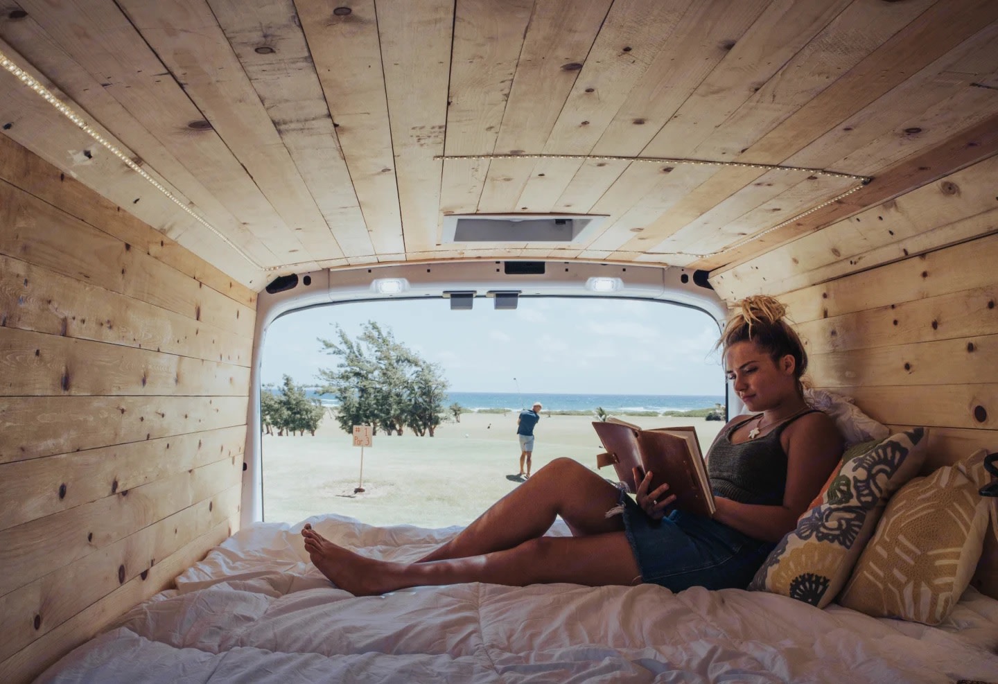 Enjoying the view and a good book from a cozy RV rental. Wooden interior, ocean, and golf course visible through the open back.. Ford Transit 2020