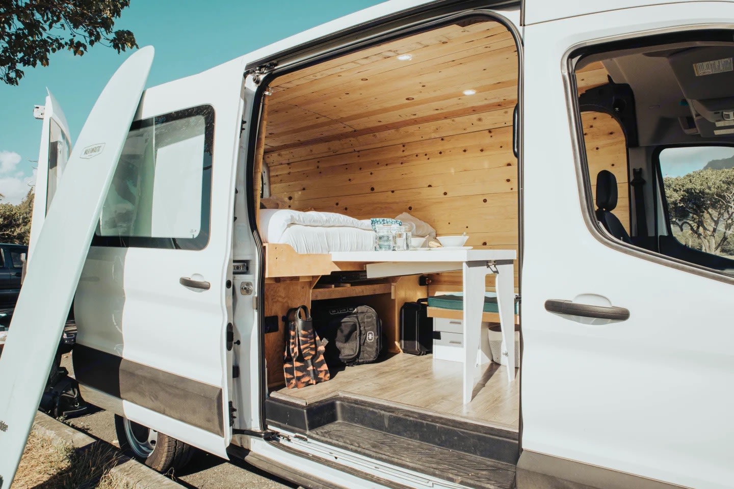 This RV rental shows a white camper van with its side door open, revealing a cozy wooden interior with a bed, table, and a surfboard outside.. Ford Transit 2020