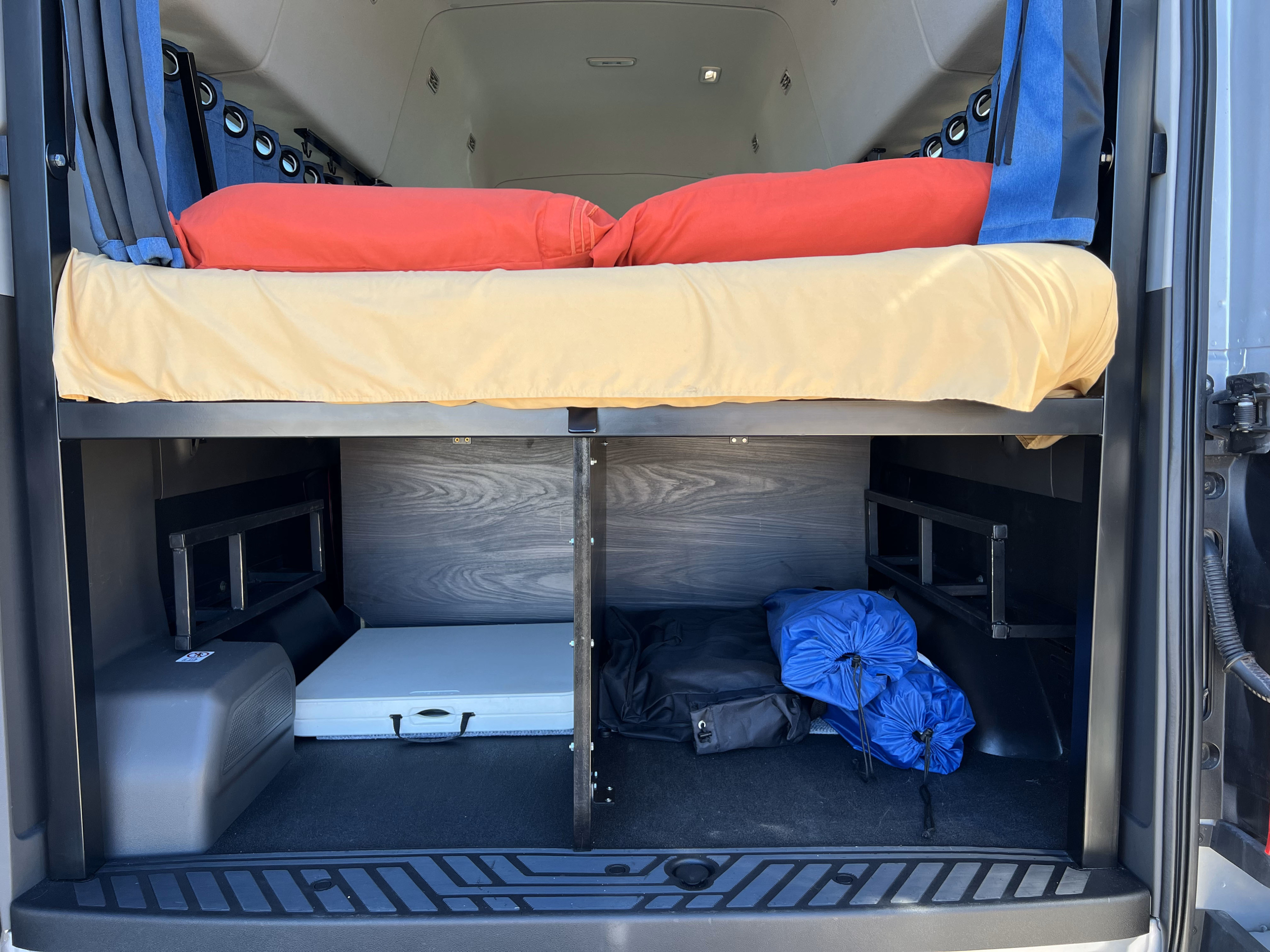 RV rental van's rear: raised bed with red pillows, blue curtains, and under-bed storage for table/bags.. Ford Transit 2019