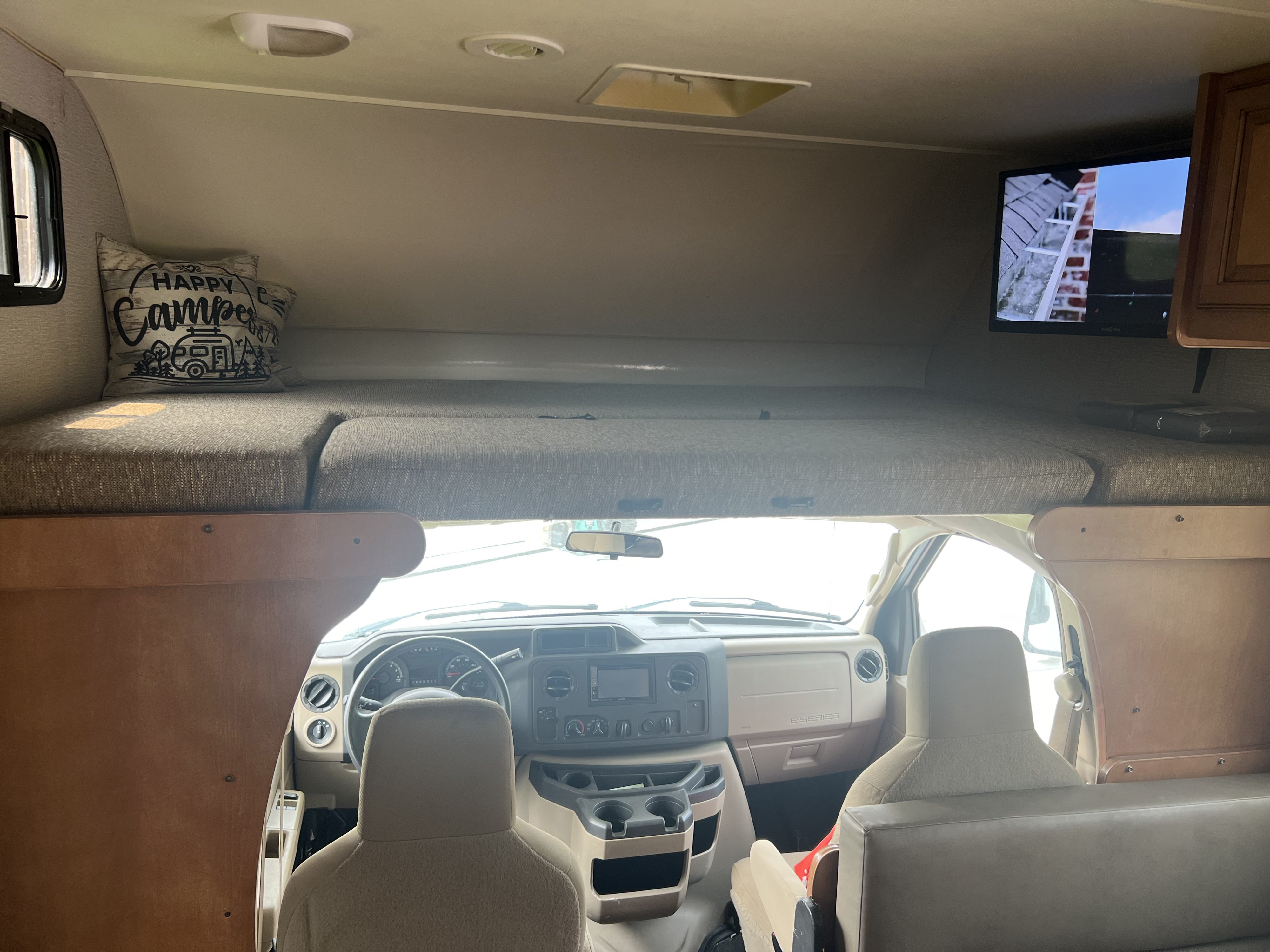 Inside a bright RV rental: cozy cab-over bunk with 'Happy Camper' pillow, TV, and driver's cockpit.. Winnebago Minnie Winnie 2015