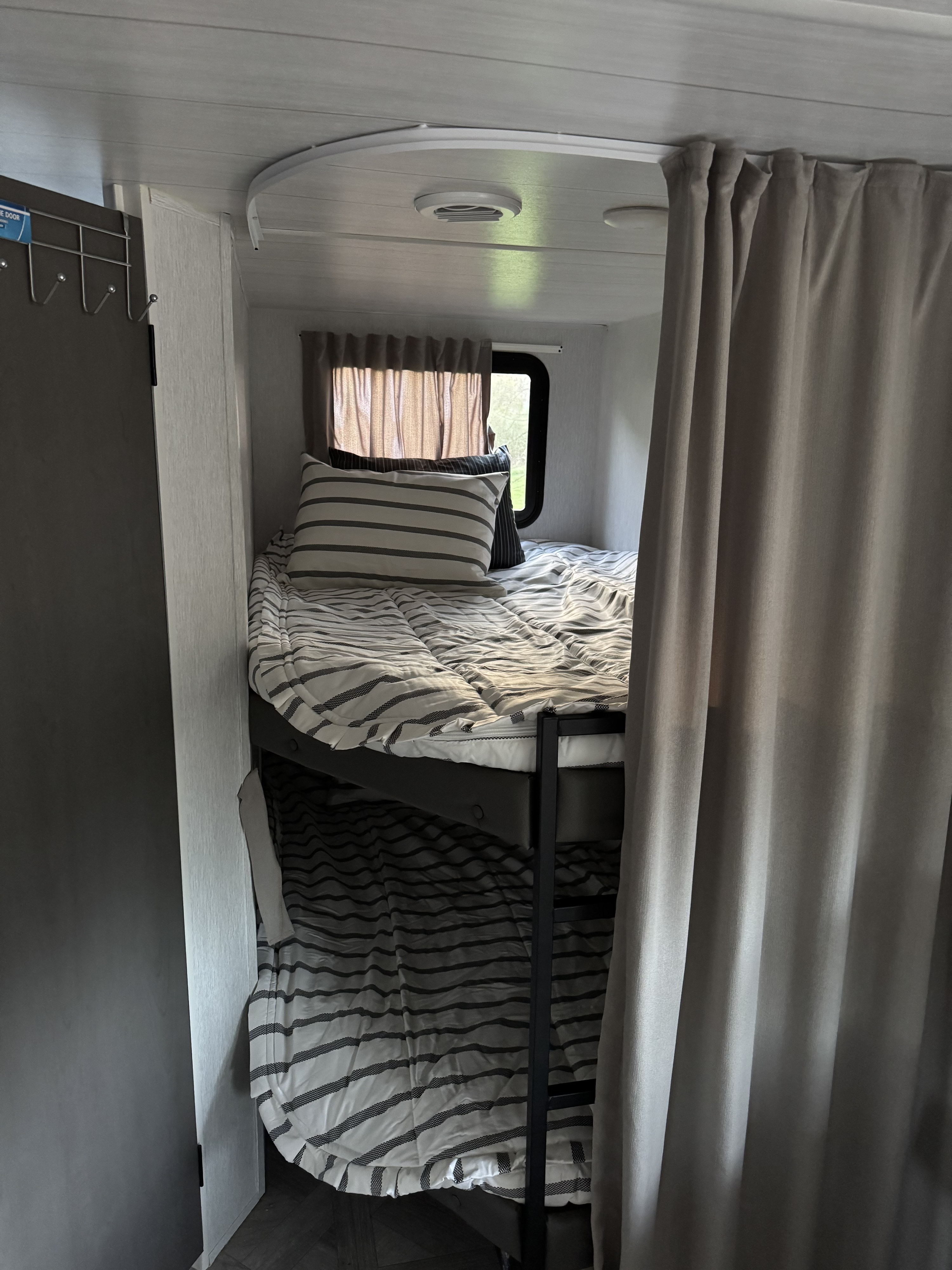 Interior view of an RV rental featuring comfortable bunk beds with striped bedding, a window, and a privacy curtain.. Forest River Wildwood X-Lite 2023