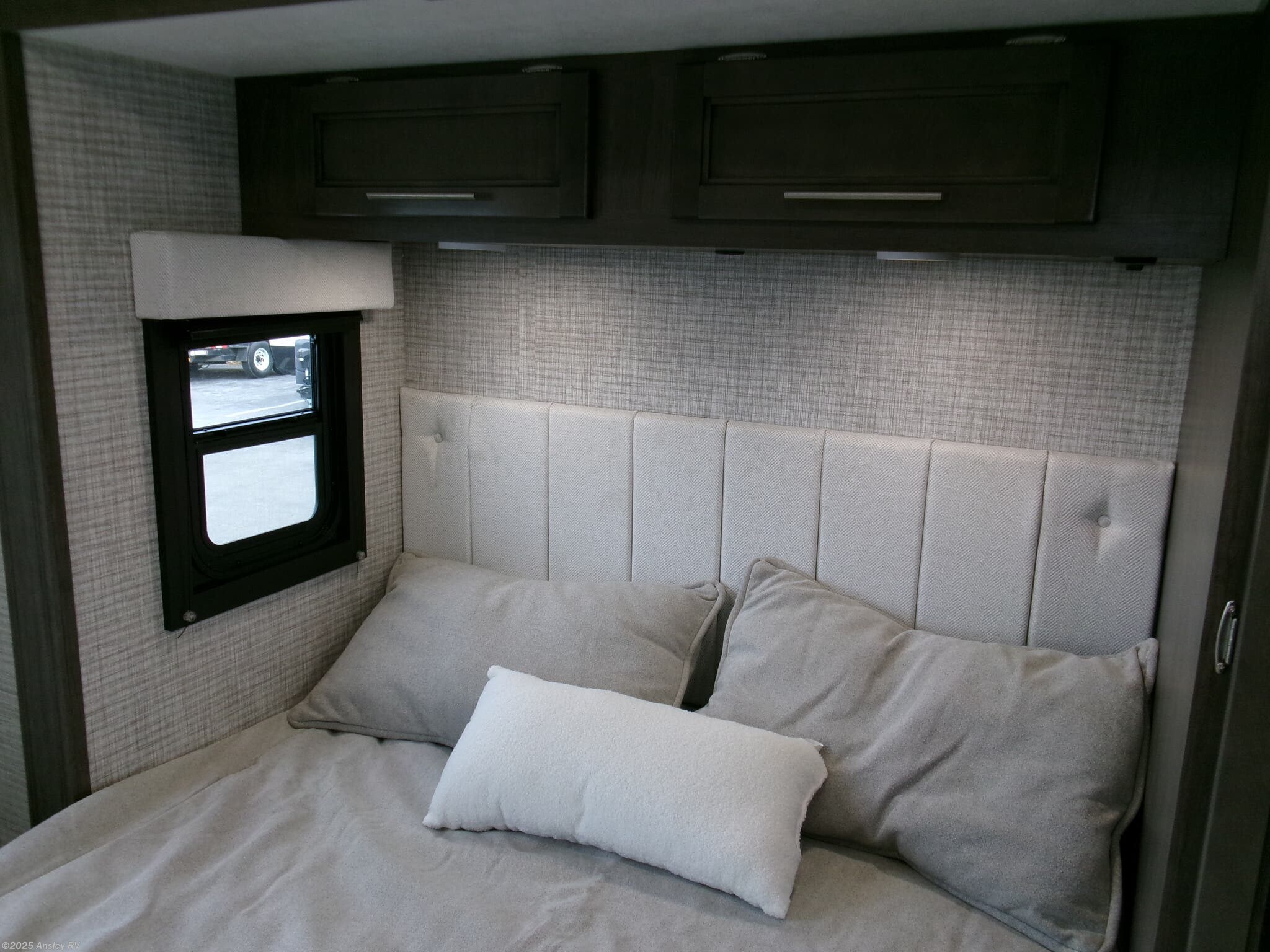 RV rental: A cozy bedroom featuring a light gray bed, upholstered headboard, window, and dark wood overhead storage.. Jayco Melbourne 2023