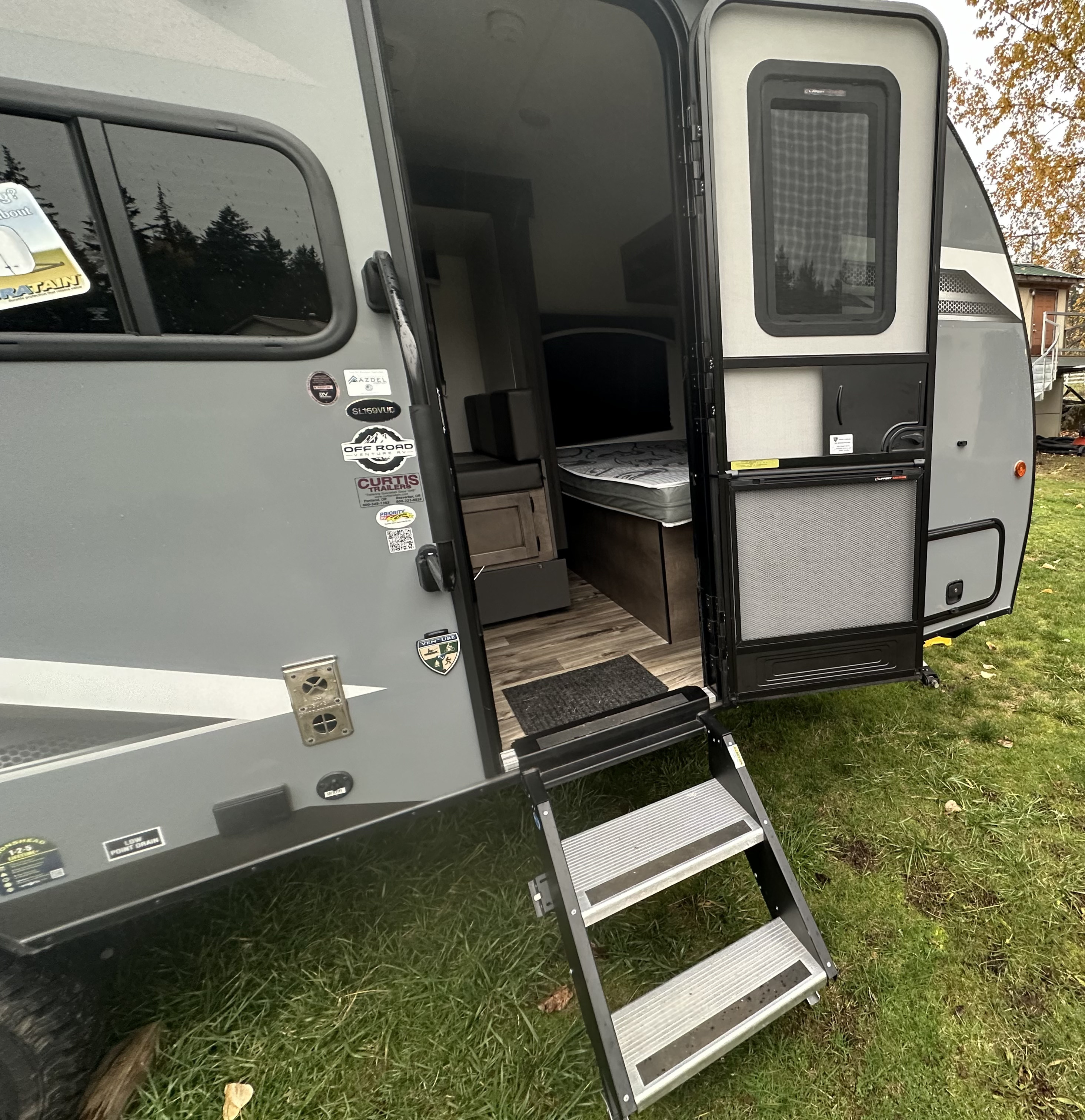 An RV rental with its side door open, revealing a cozy interior with a bed. Extended steps lead to the grassy ground.. Venture RV SL169VUD 2023
