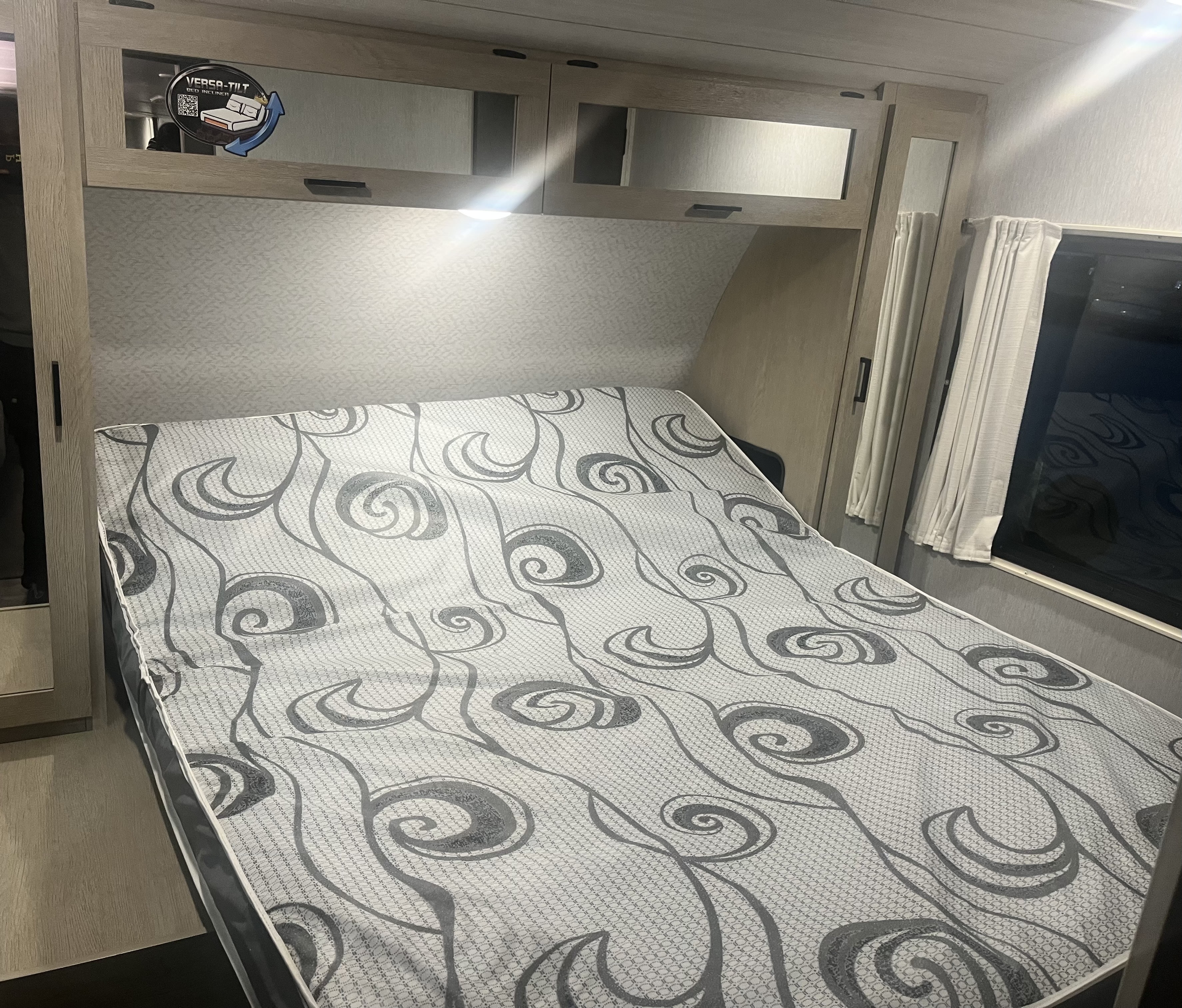 Cozy RV rental bedroom featuring a 'VERSA-TILT' bed, patterned mattress, overhead cabinets, and window with curtains.. Forest River Wildwood Platinum 2024
