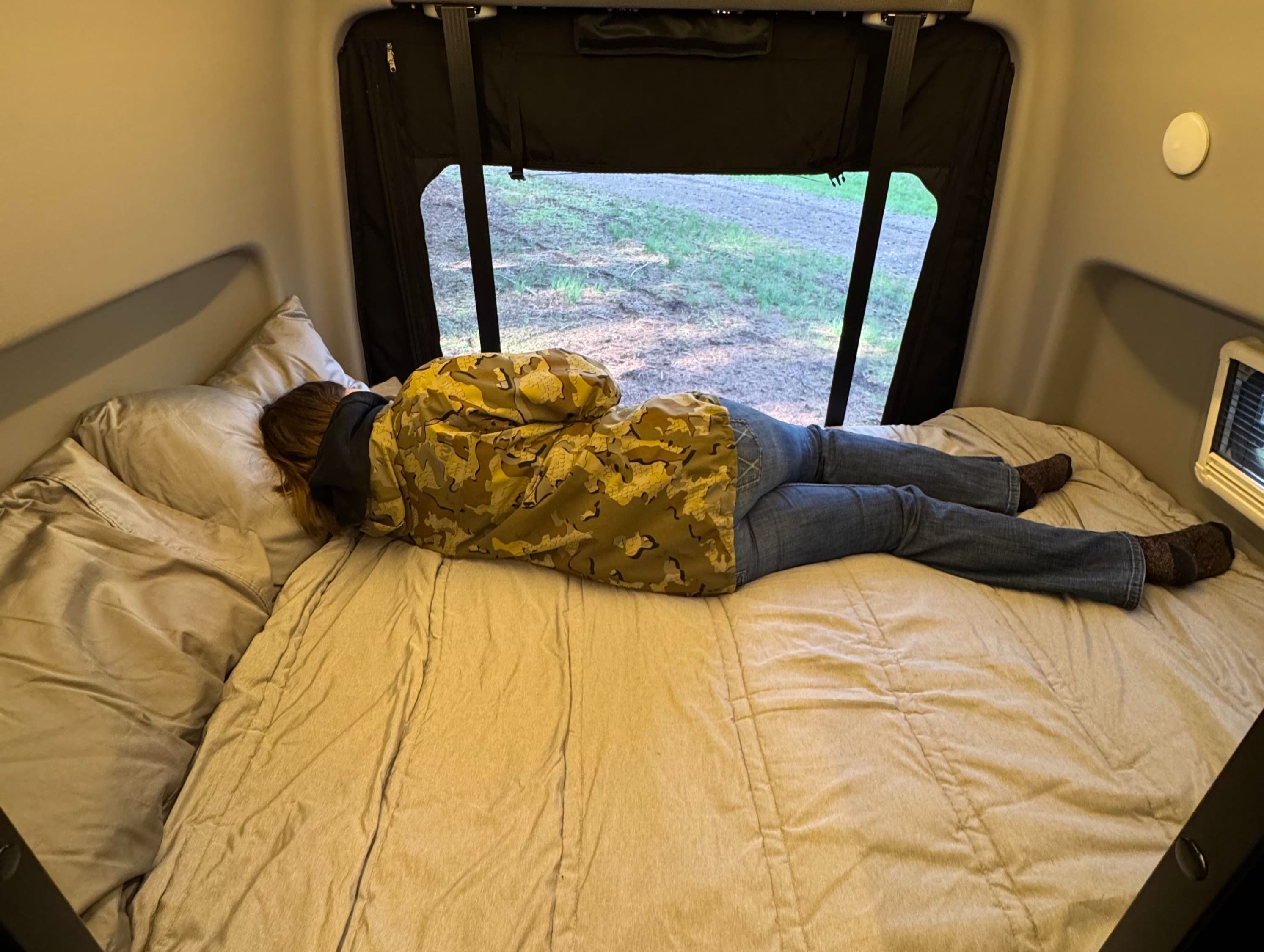 A person in camo and jeans relaxes face down on a bed in an RV rental, looking out the window.. Winnebago Revel 2020