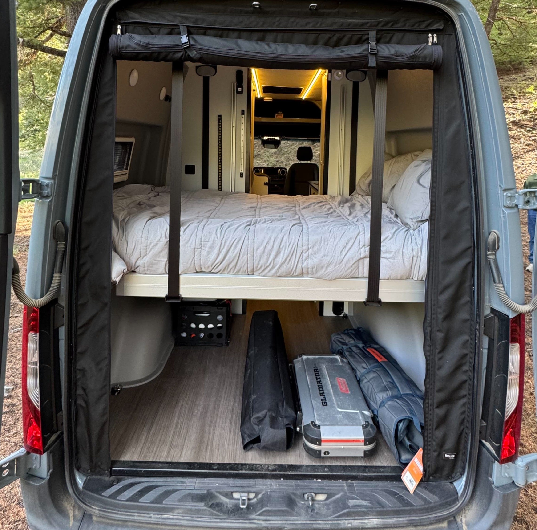 RV rental: Inside a camper van, showing a bed over under-bed storage on a wood floor, with open rear doors.. Winnebago Revel 2020