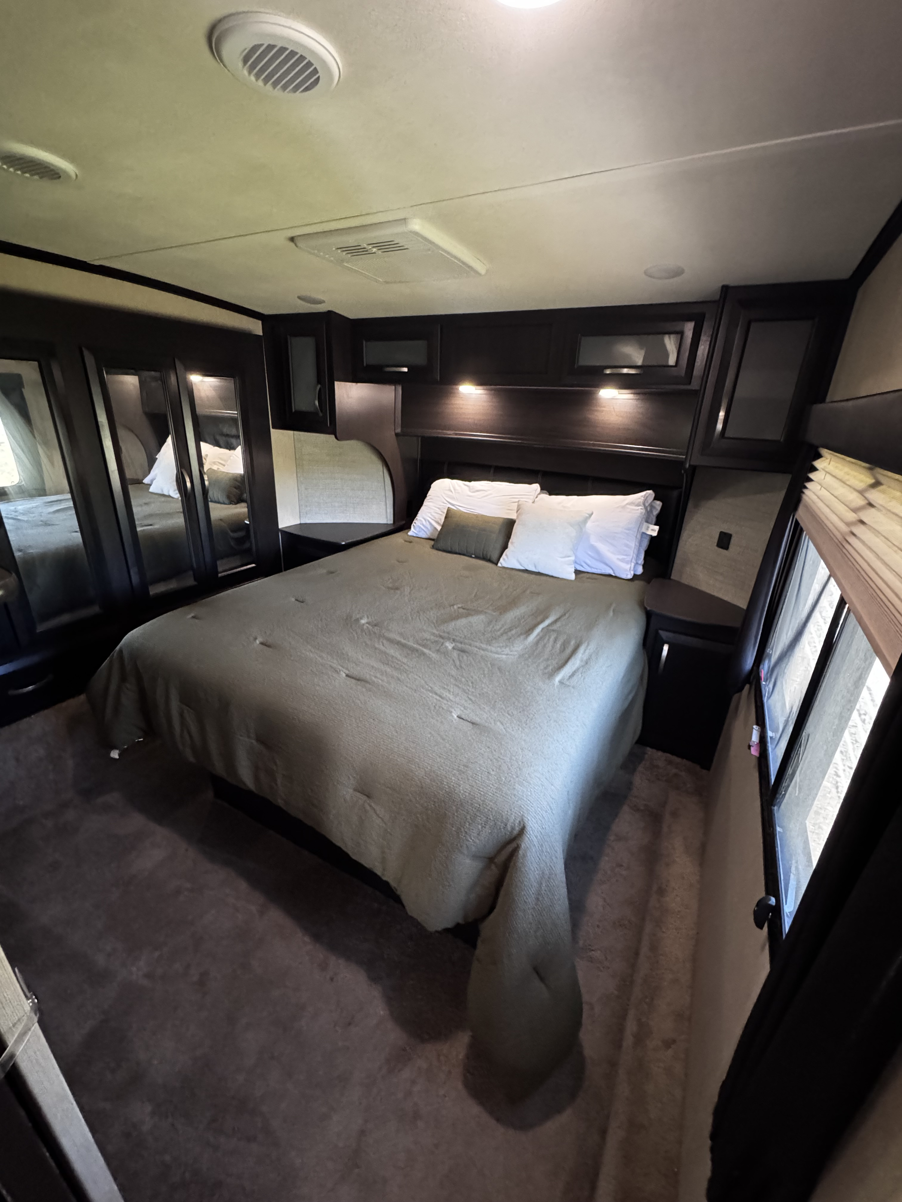 This image shows the compact yet comfortable bedroom interior of an RV rental, featuring a bed, built-in cabinetry, and ceiling vents.. Dutchmen Voltage 2018