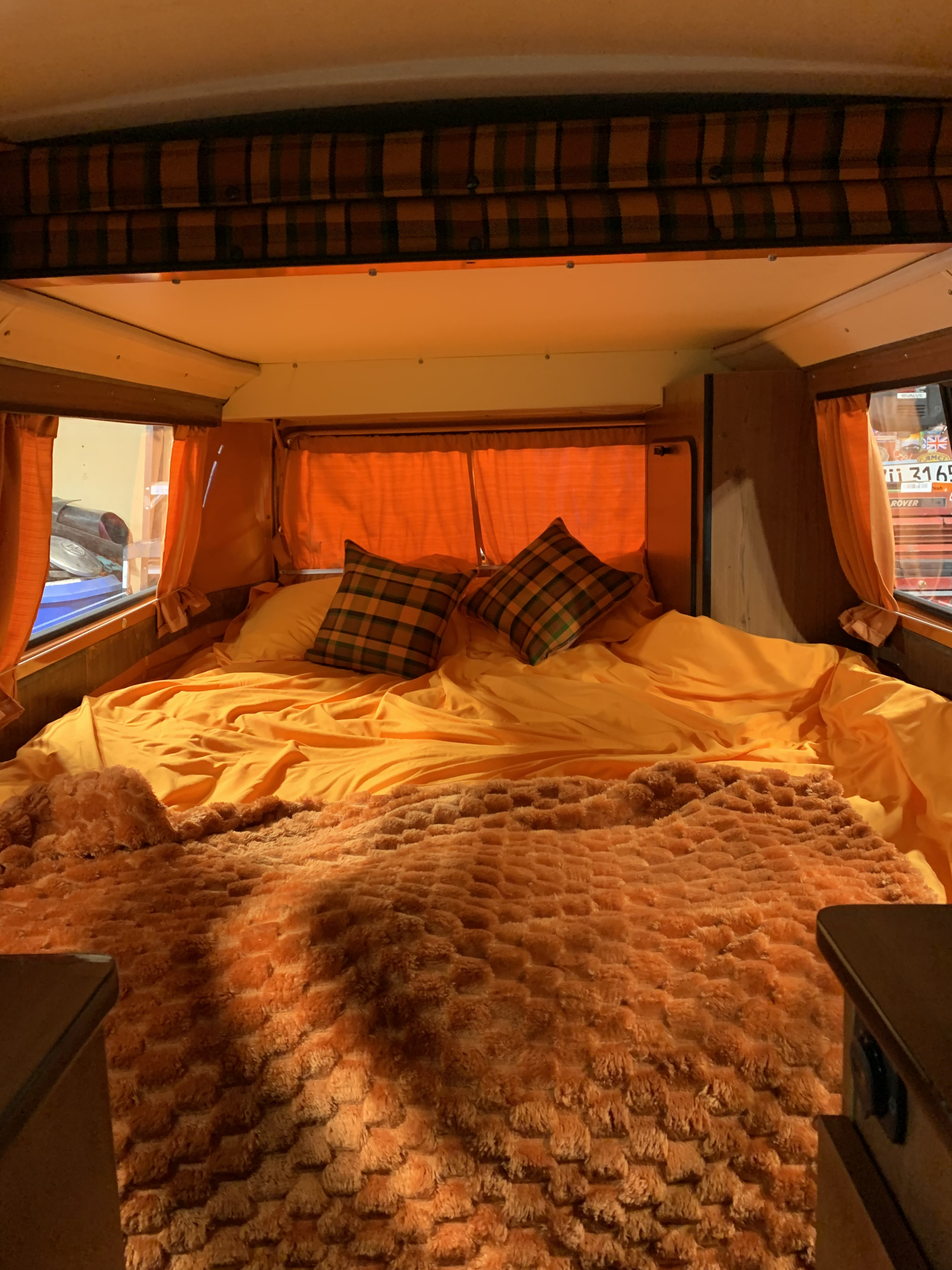 Vibrant RV rental bed setup: bright orange sheets, a plush, textured orange blanket, and two plaid pillows inside a cozy camper.. Volkswagen Kombi T2 Westfalia 1974