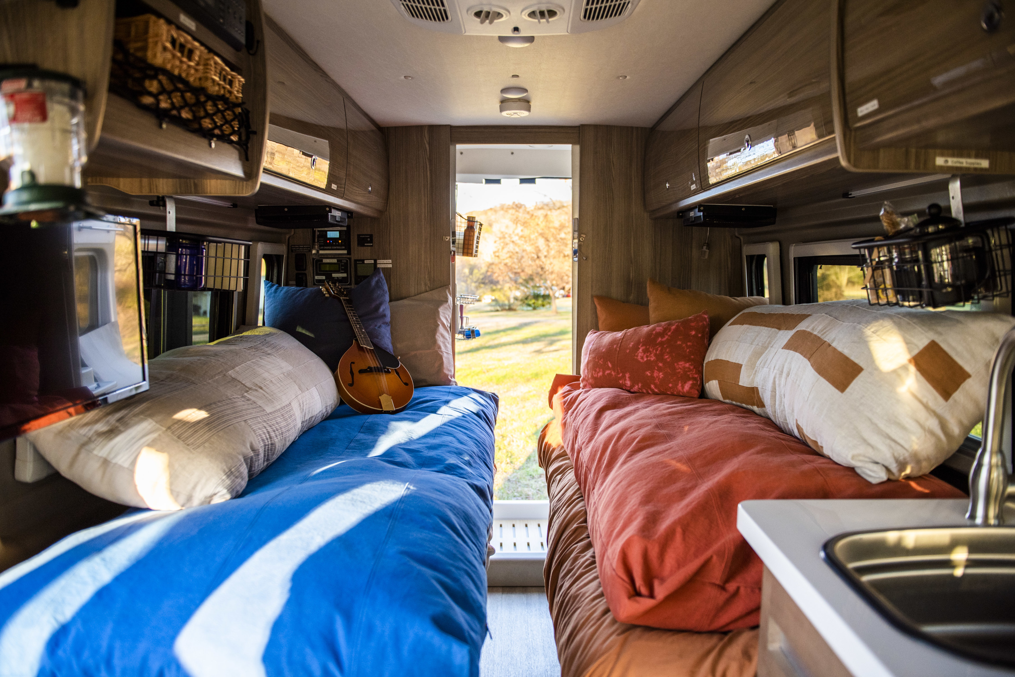 Bright RV rental interior: two cozy beds, one with a mandolin, overhead storage, and a sink, overlooking a scenic outdoor view.. Winnebago Travato 2020