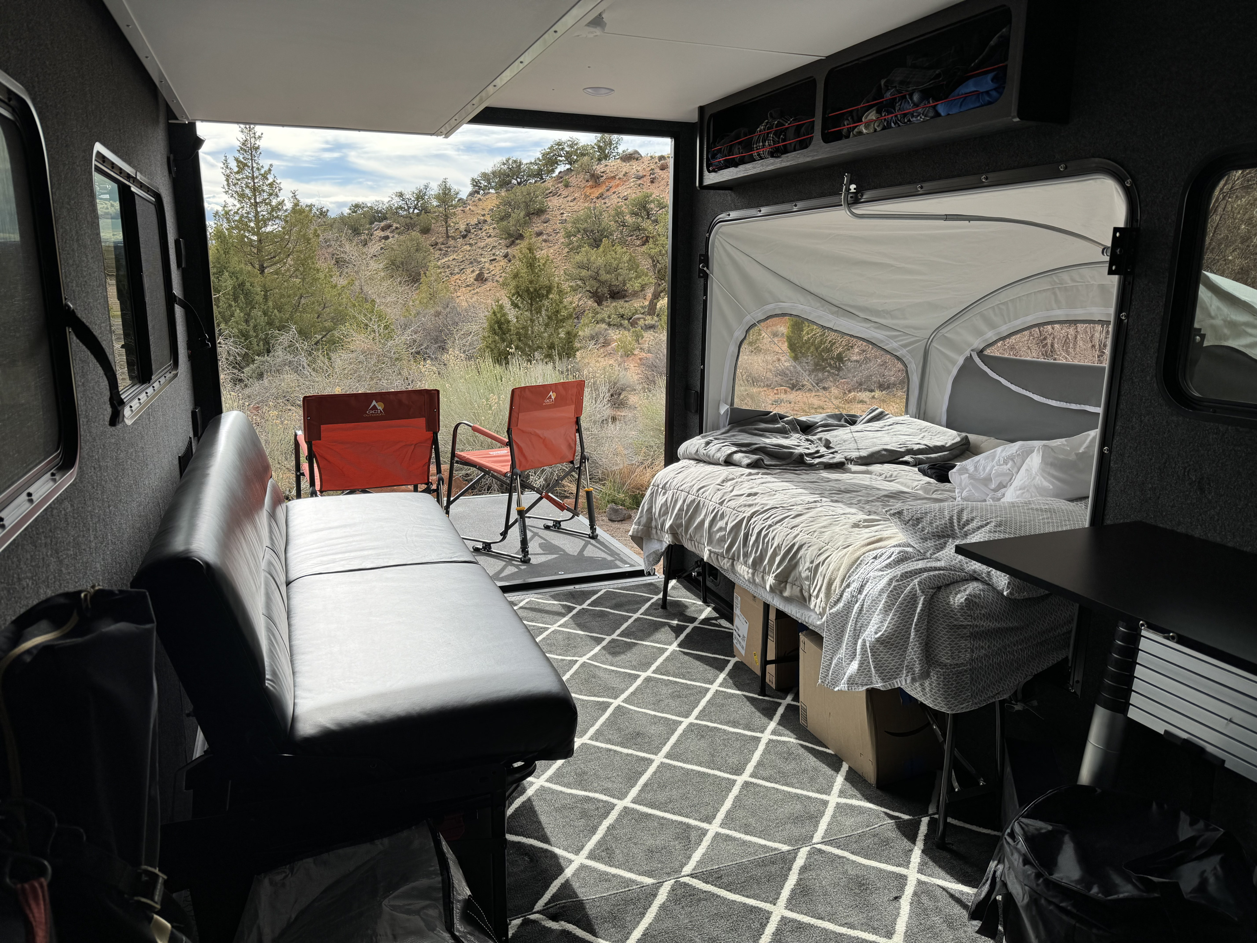 Cozy RV rental interior with bed, long black bench seat, and a rear ramp patio with chairs opening to a desert view.. InTech RV Forge 2024