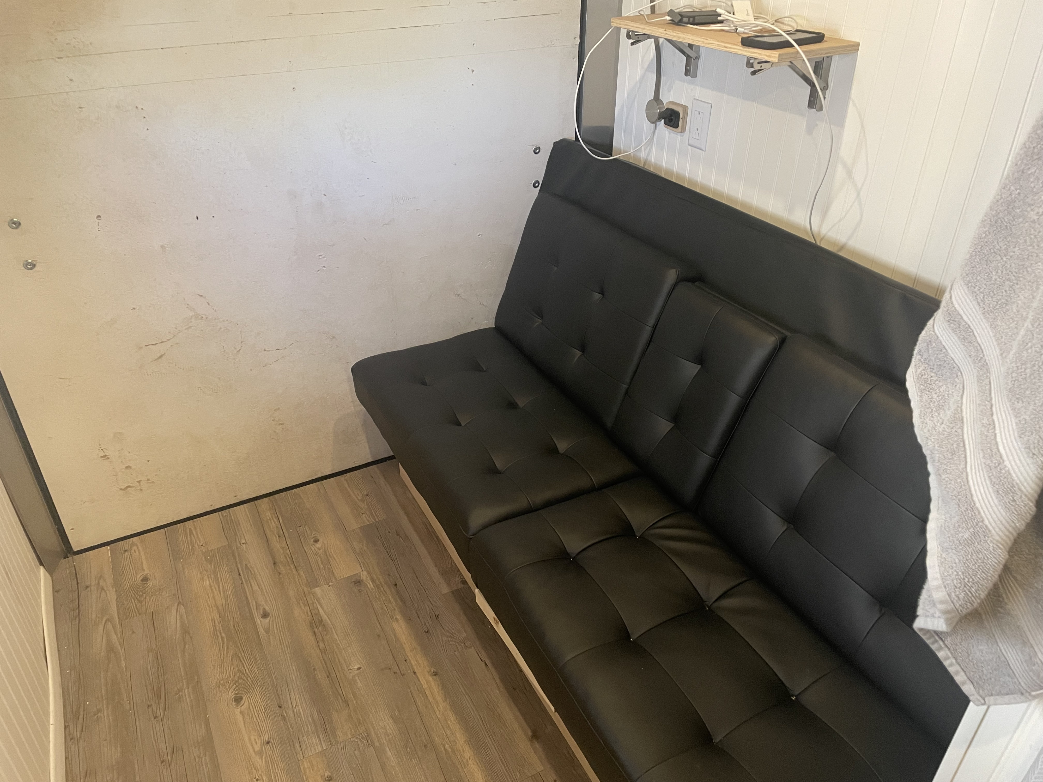 Cozy RV rental interior featuring a black futon, charging shelf, and wood-look flooring.. Wells Cargo 6x12 enclosed conversion travel 2023