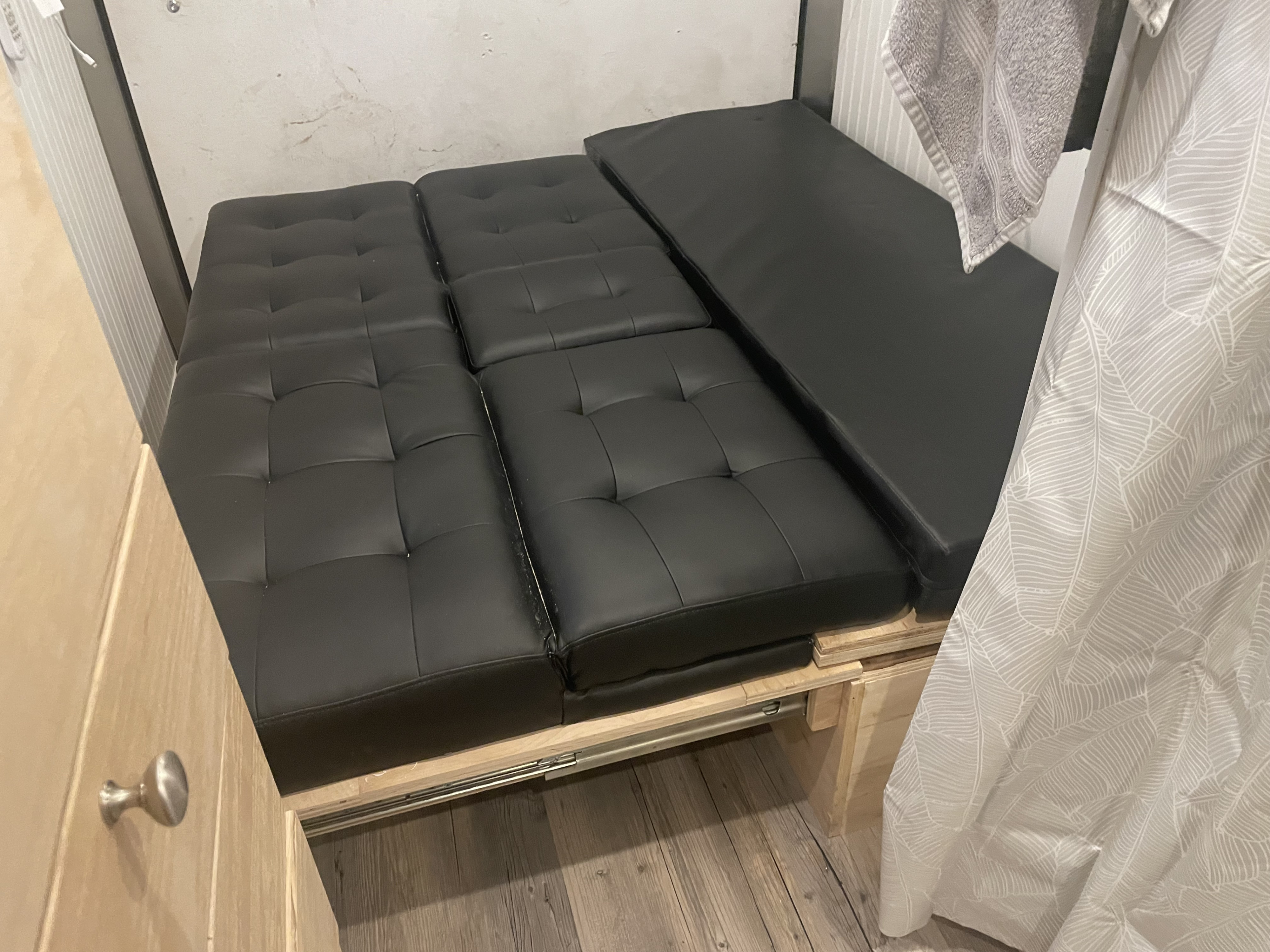 RV rental: A compact interior features black tufted cushions forming a pull-out bed system on wooden rails, maximizing space.. Wells Cargo 6x12 enclosed conversion travel 2023
