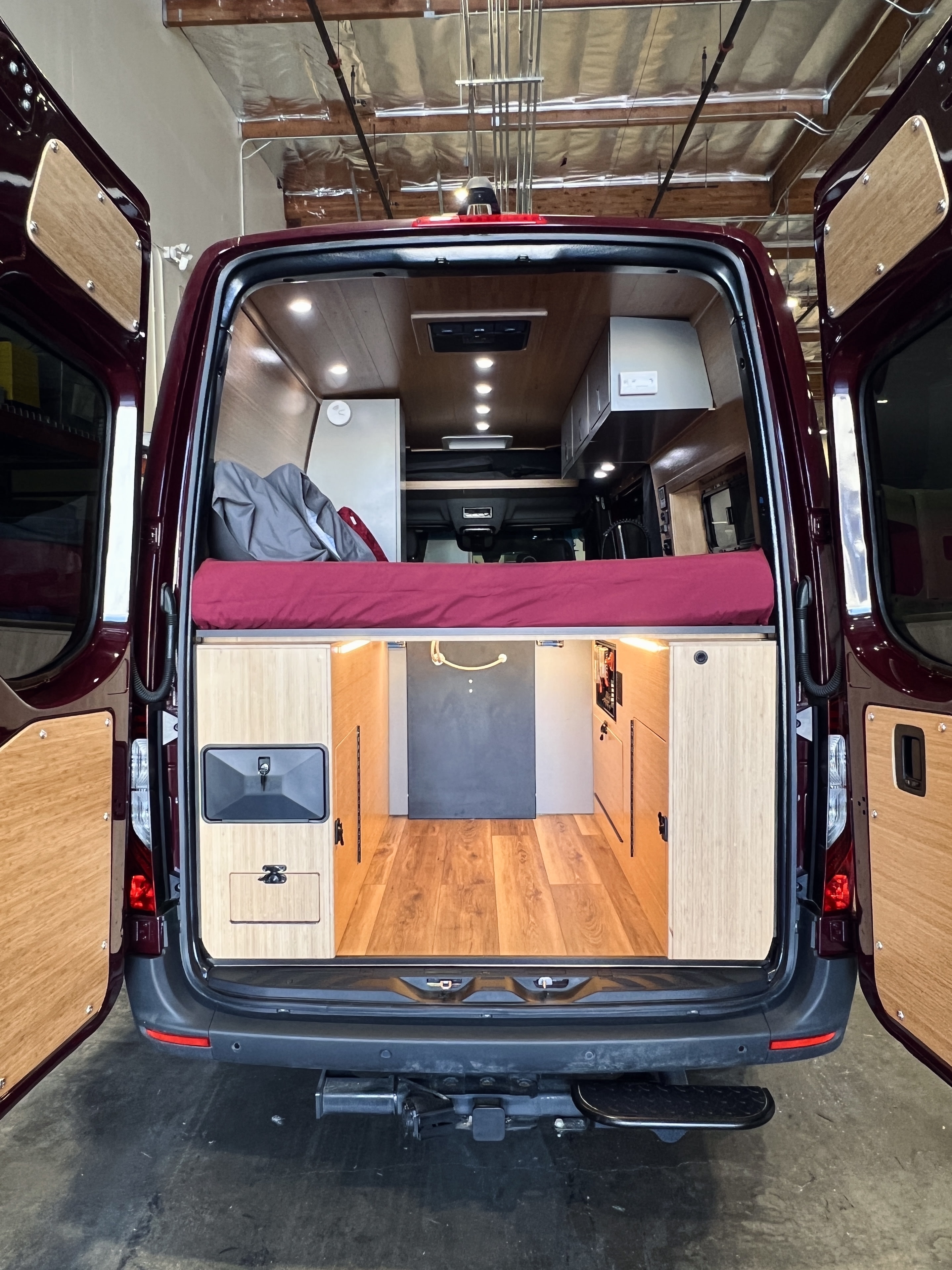 Rear view of an RV rental camper van with open doors, showing a bed, wood floor, and custom storage.. Mercedes-Benz Sprinter 2025