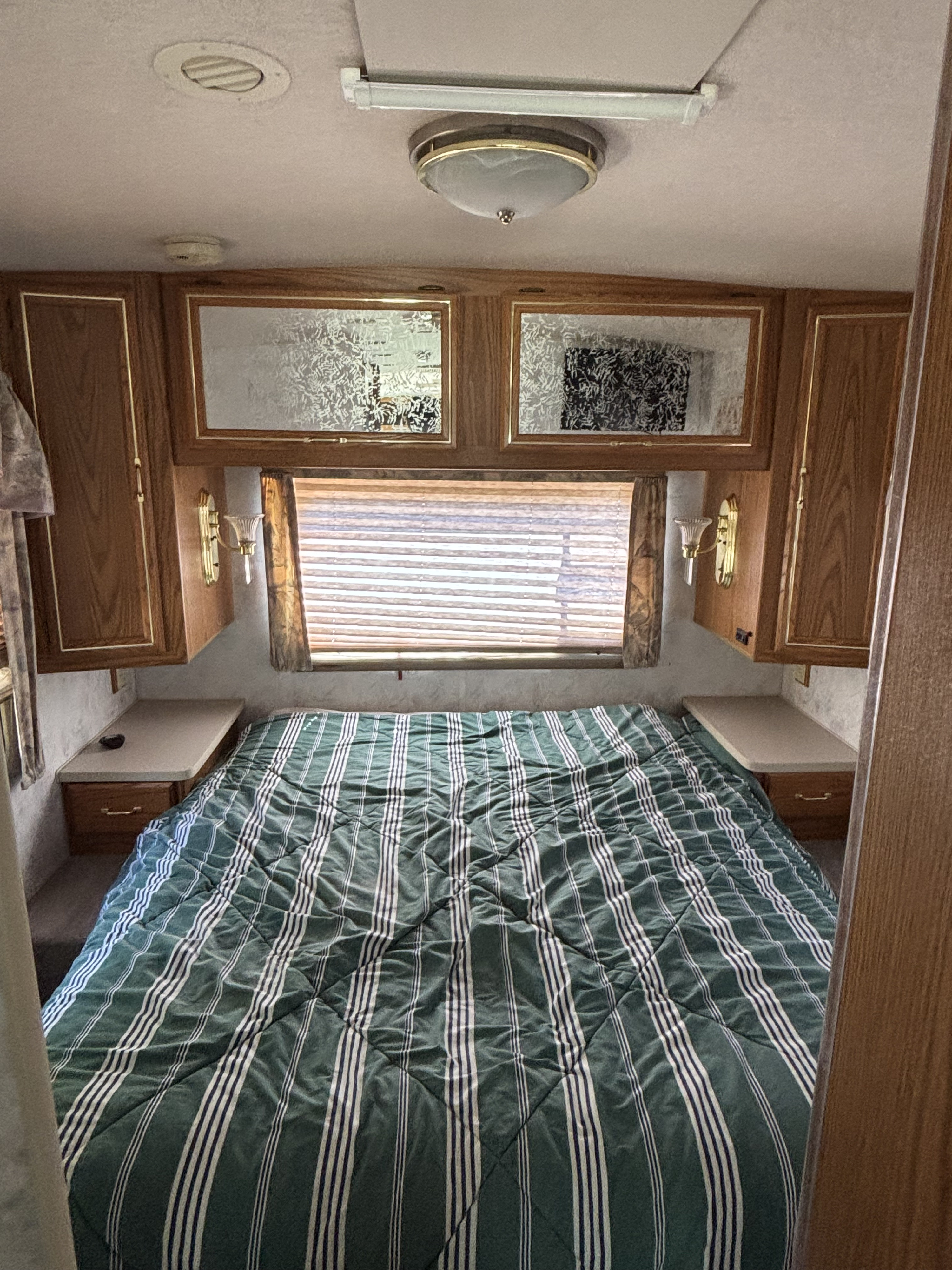 Cozy RV rental bedroom featuring a green striped bed, built-in wood cabinets, and a window with blinds.. Winnebago Brave 1997