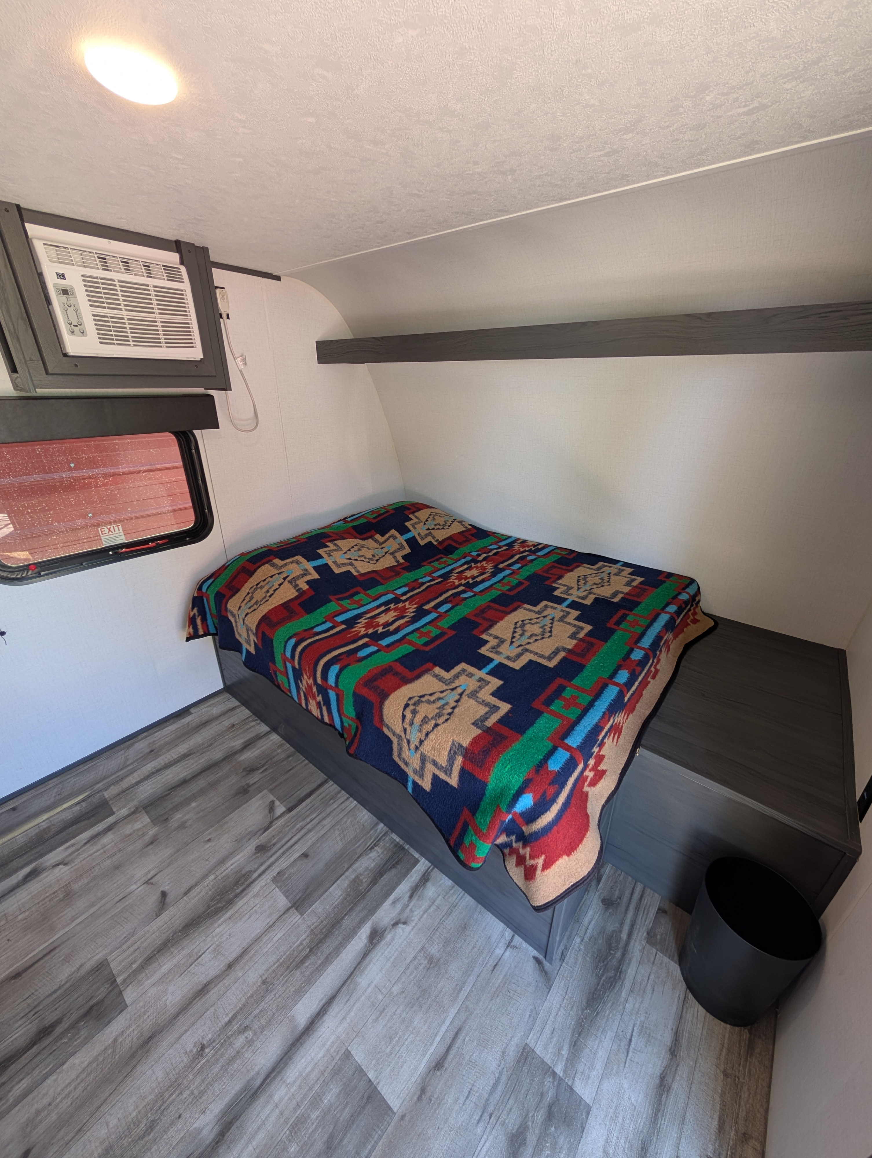 Inside an RV rental, a compact bedroom features a colorful patterned bed, an air conditioner, and wood-look flooring.. Aspen Trail 17RB 2024