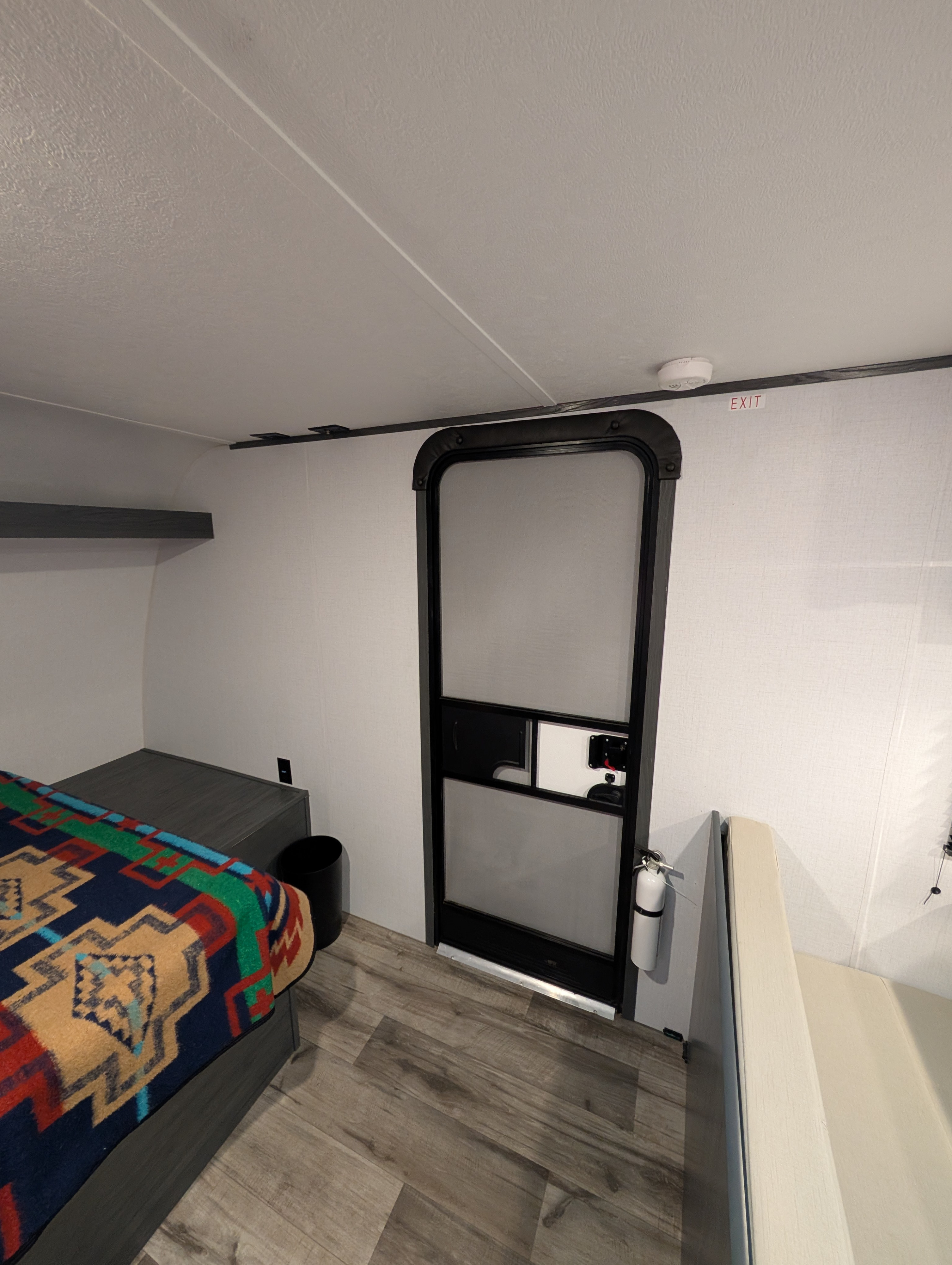RV rental interior showcasing a cozy bed, main entry door with fire extinguisher, exit sign, and textured flooring.. Aspen Trail 17RB 2024